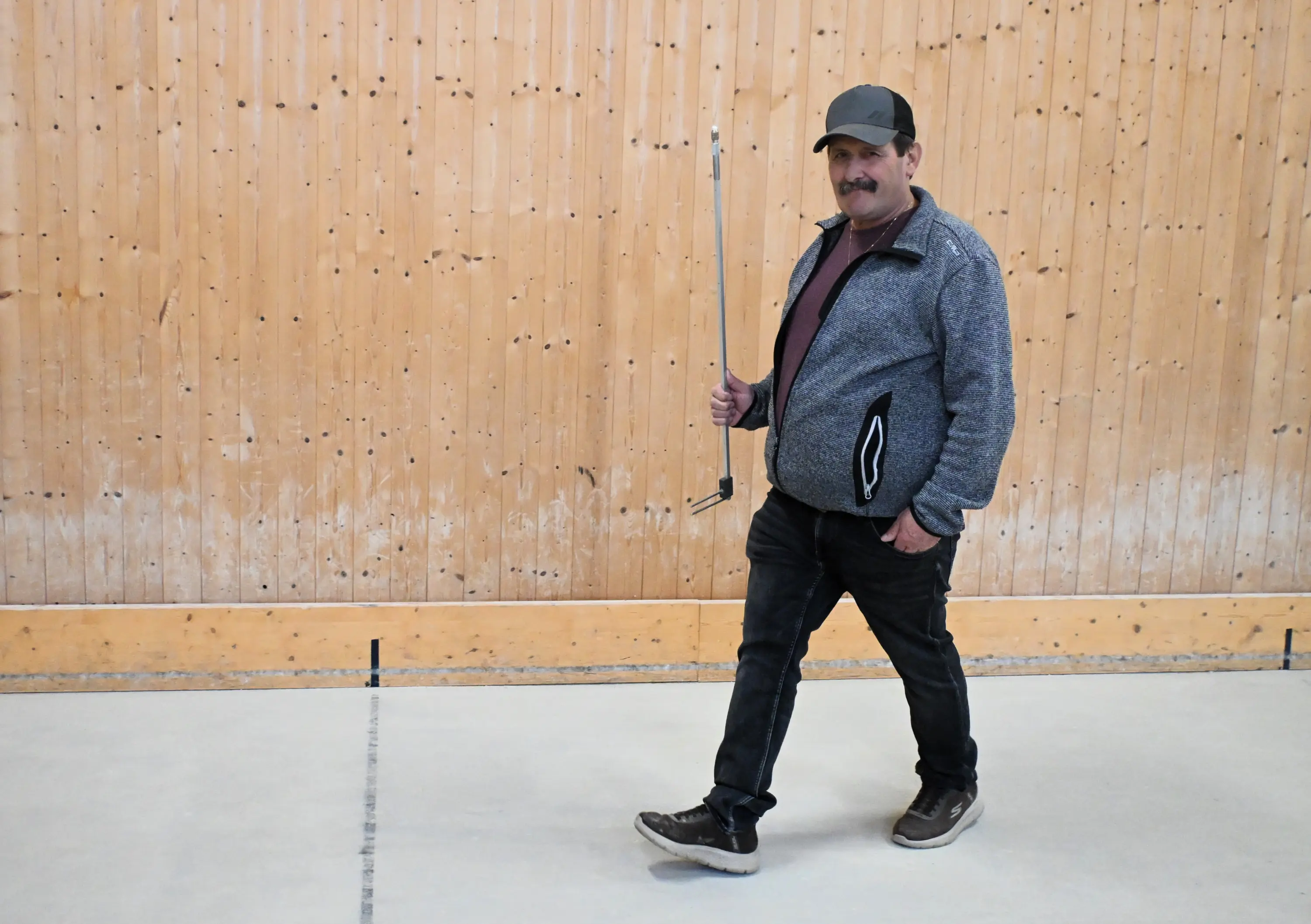 Uomo che cammina all'interno di una sala con pareti in legno. Indossa una giacca grigia e un cappello nero, tenendo in mano una barra di metallo.