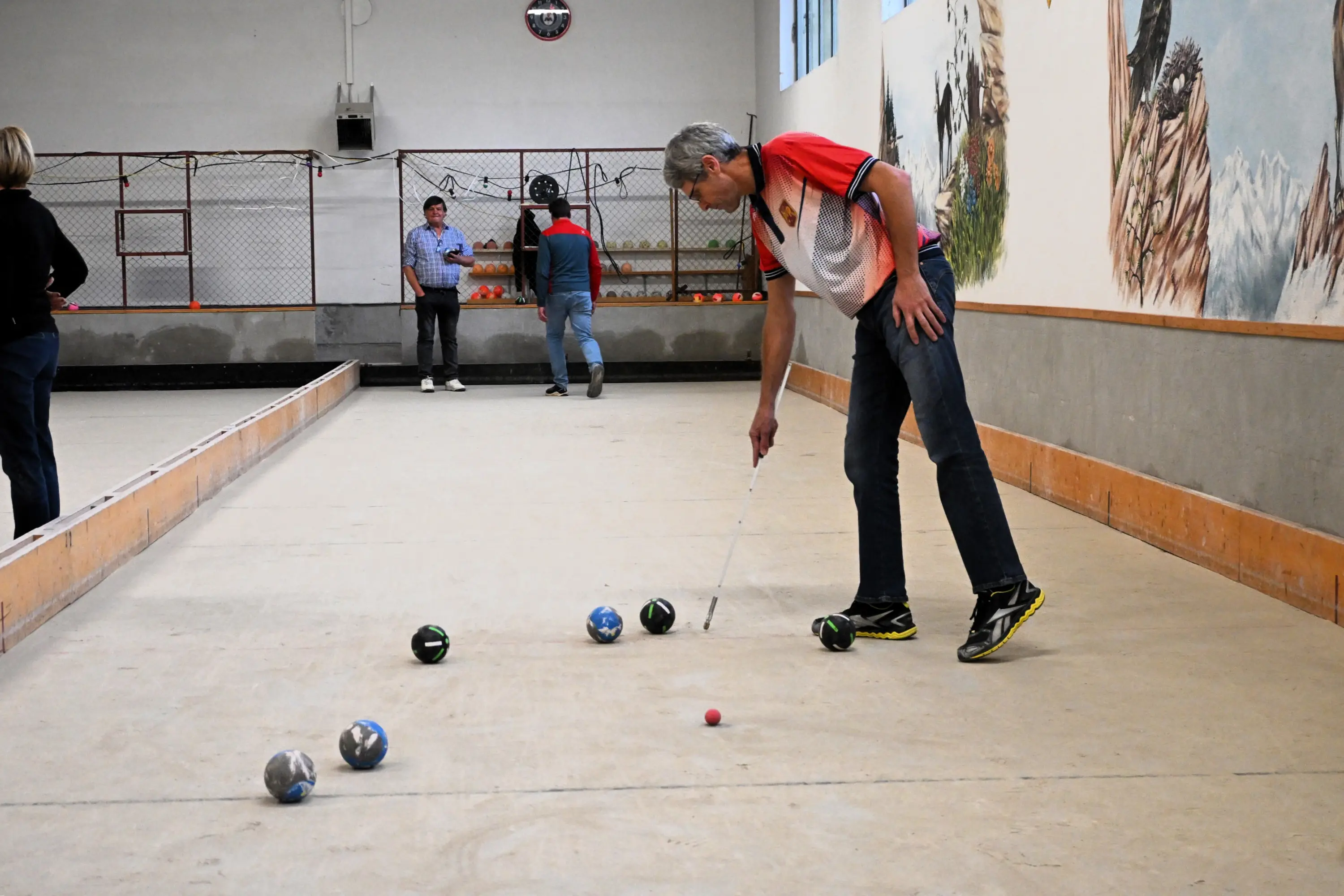 Un uomo si prepara a tirare una pallina in un campo da bocce. Alcuni giocatori si trovano sullo sfondo, mentre il muro è decorato con un grande murale.