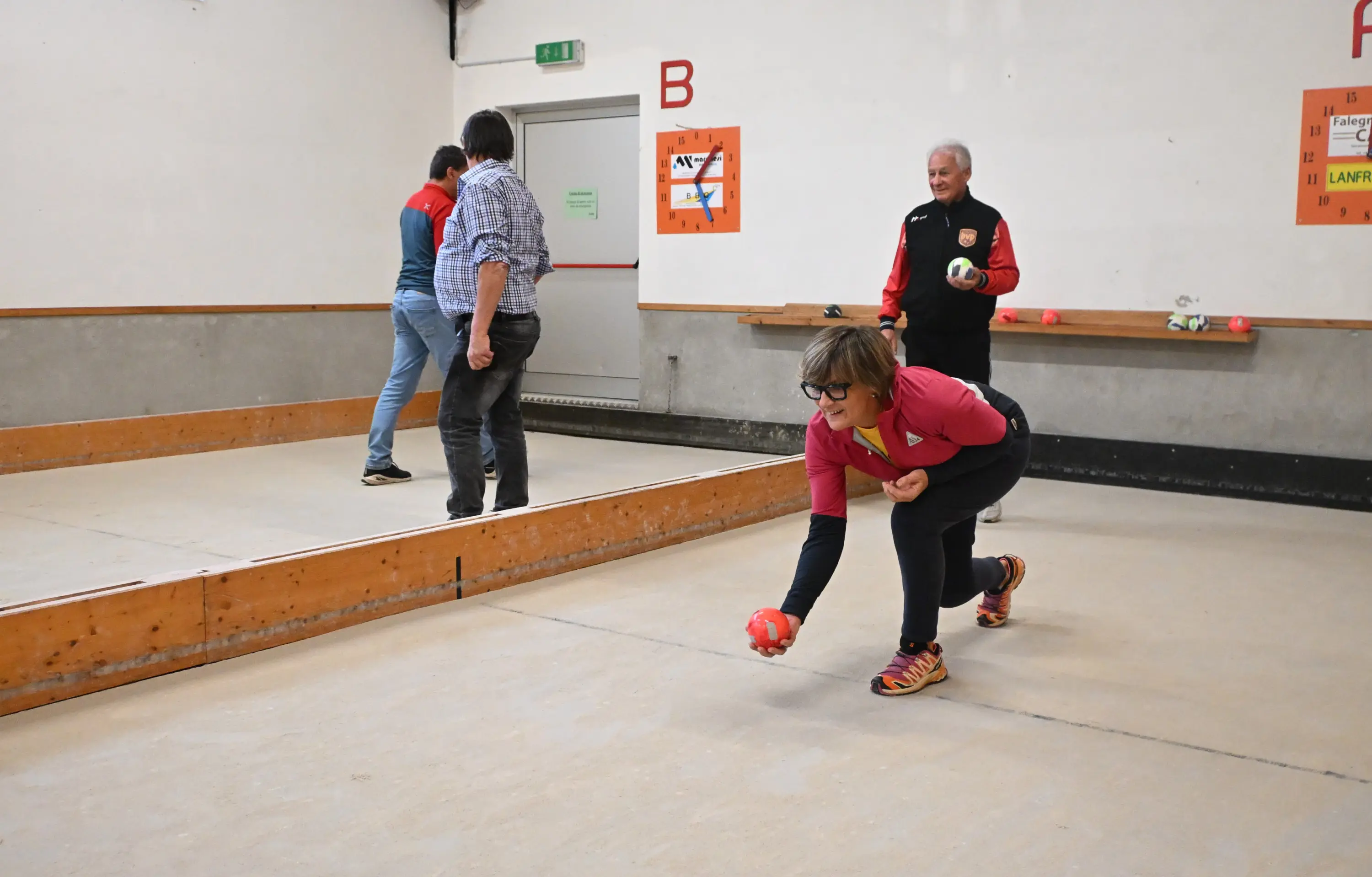 Una donna in maglietta rossa lancia una boccia in una sala da gioco. Due uomini sono visibili sullo sfondo, uno in piedi e l'altro mentre si prepara a lanciare. Le pareti sono decorate con numeri, e il pavimento è in cemento
