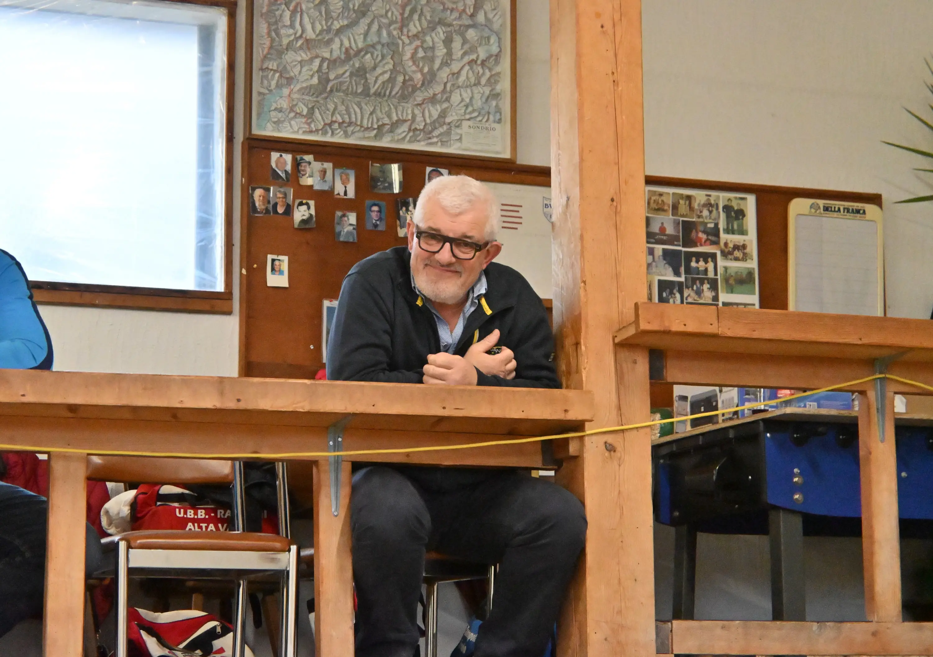 Un uomo con occhiali e una felpa scura sorride mentre si trova seduto a una scrivania in un ambiente interno, circondato da fotografie appese al muro.