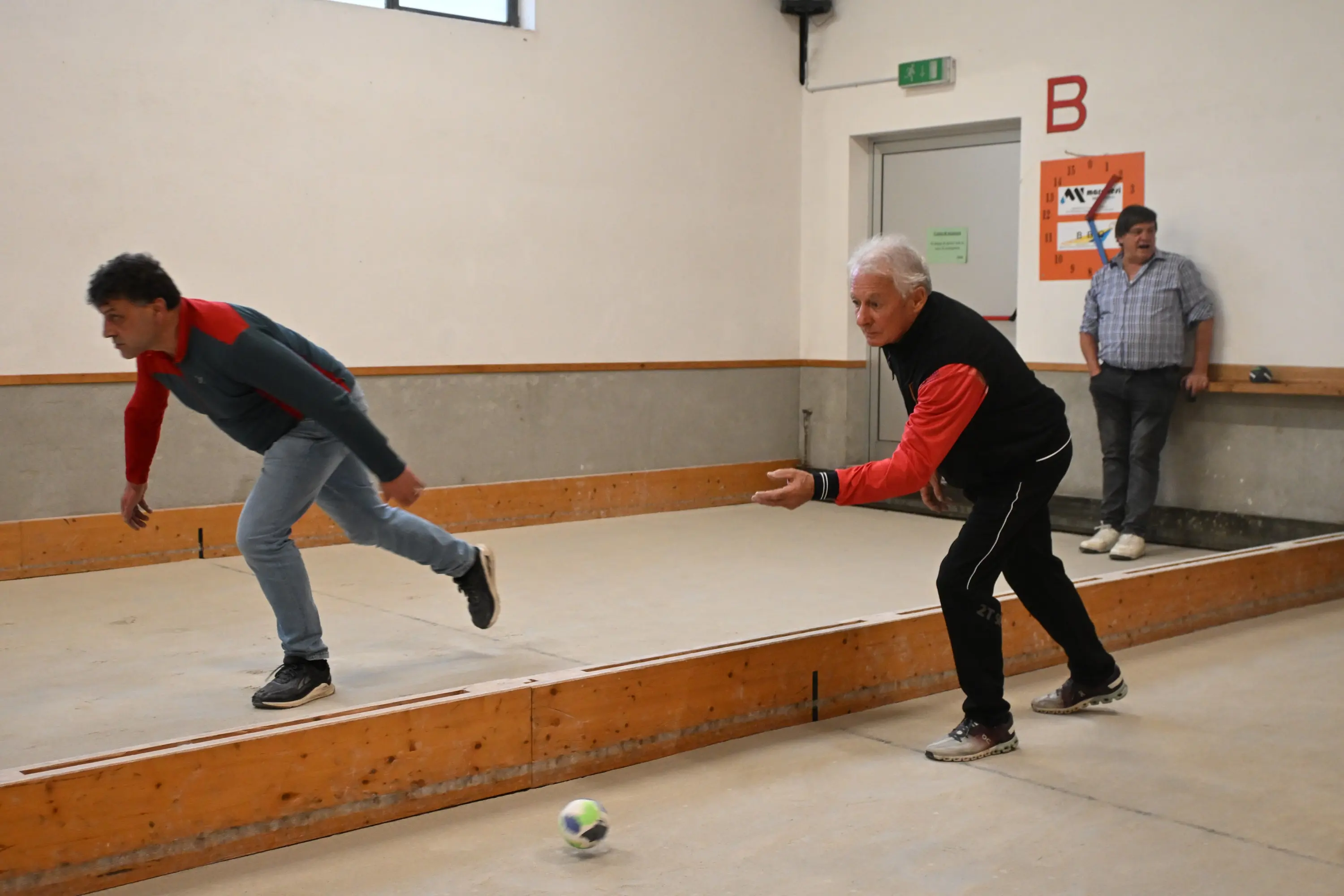 Due uomini stanno giocando a bocce in una sala. Un uomo si appresta a lanciare la boccia mentre l'altro osserva attentamente. Sullo sfondo si vede un terzo uomo che guarda la partita.
