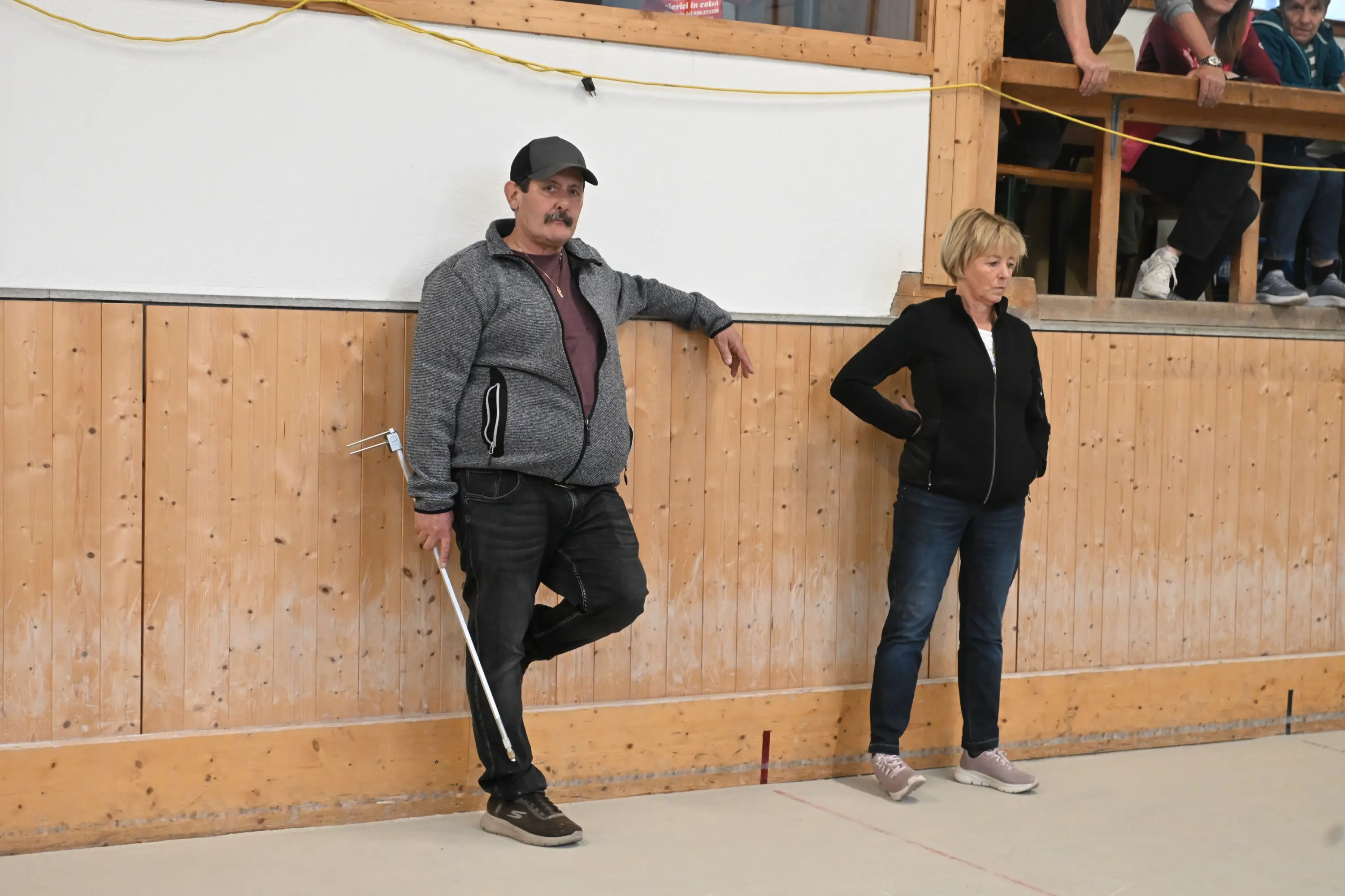 Due persone in piedi all'interno di un'arena. L'uomo indossa un cappello e una giacca grigia, mentre la donna ha i capelli biondi e indossa un giubbotto nero. Sullo sfondo si intravede un gruppo di persone sedute