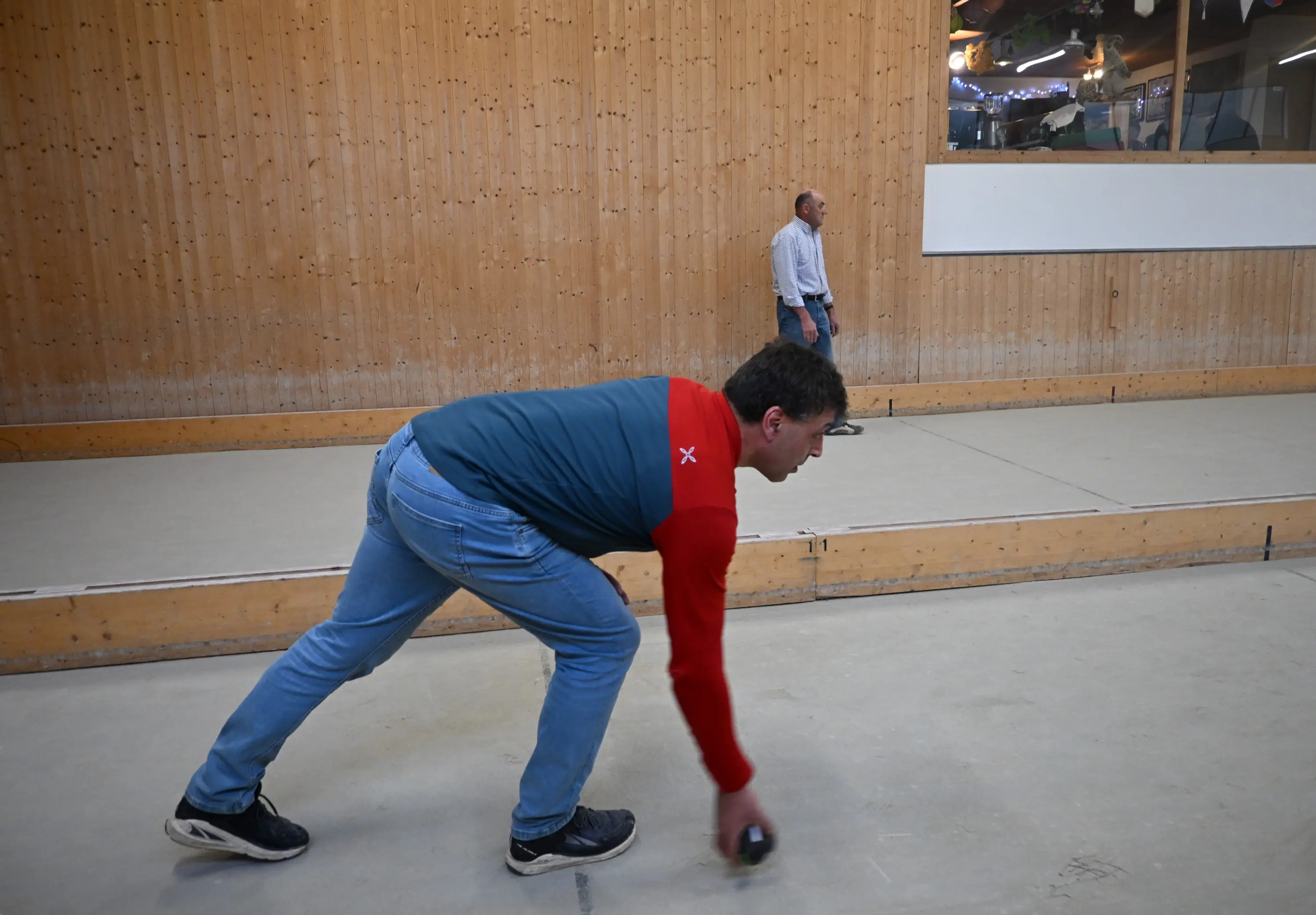 Un uomo sta lanciando una boccia all'interno di una sala con pareti di legno. Sullo sfondo, un'altra persona osserva.