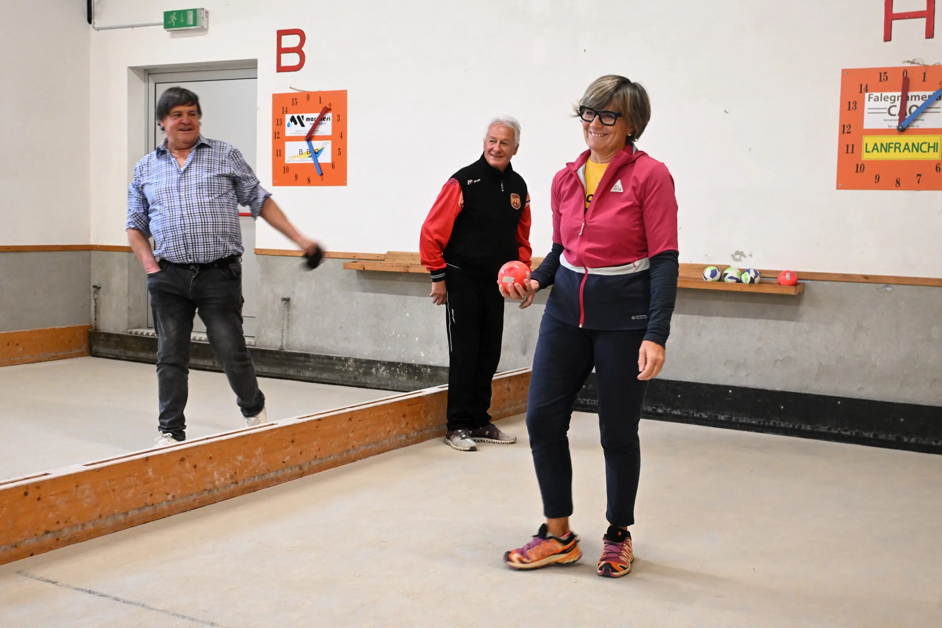 Un uomo lancia una palla in un campo di bocce mentre una donna sorridente, con occhiali e abbigliamento sportivo, si prepara a tirare. Un altro uomo, in background, osserva con interesse. Sul muro ci sono poster informativi sul gioco.