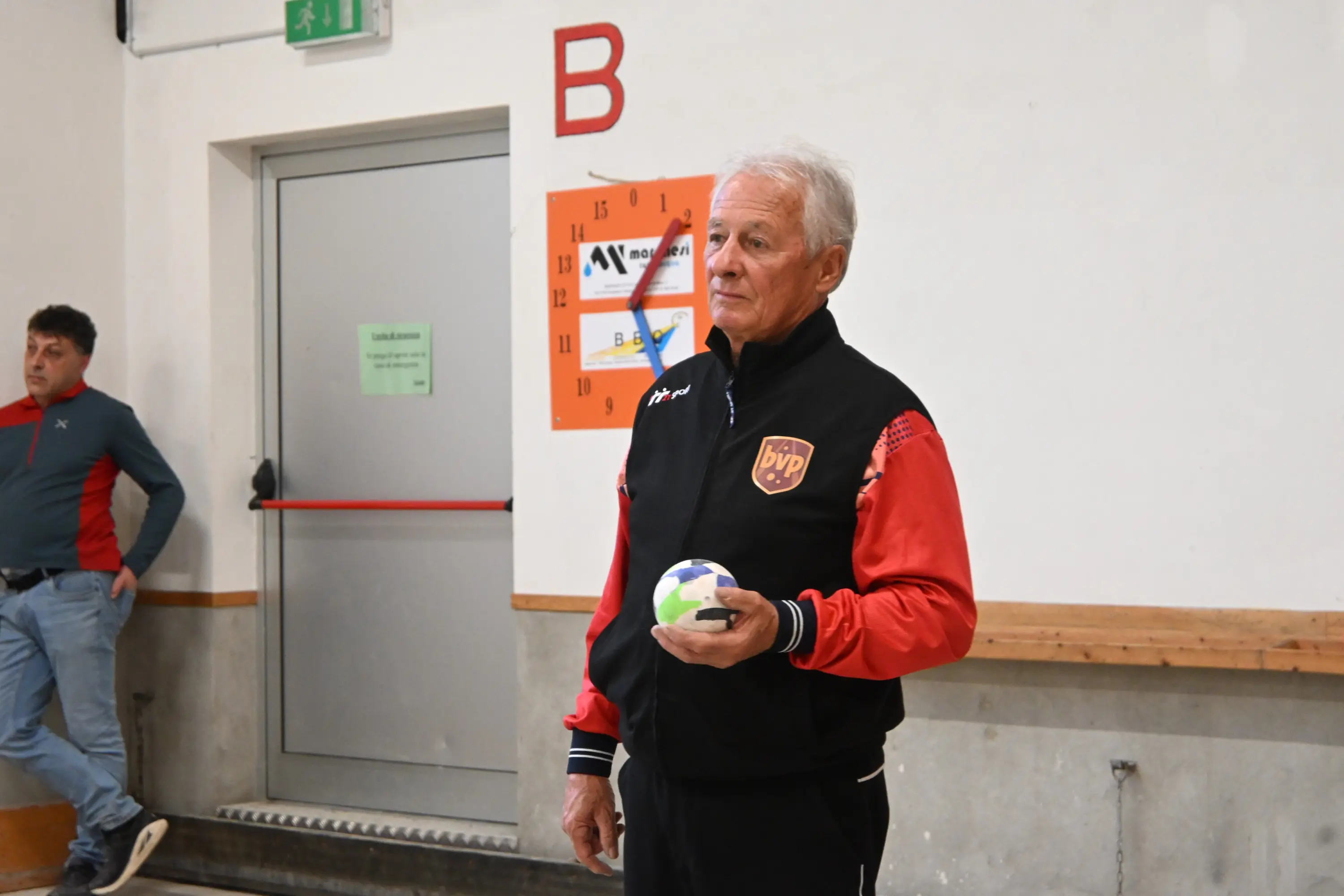 Un uomo di mezza età in giacca sportiva nera con dettagli rossi tiene una palla in mano. Sullo sfondo, un altro uomo è in piedi vicino a una porta e un calendario appeso al muro.