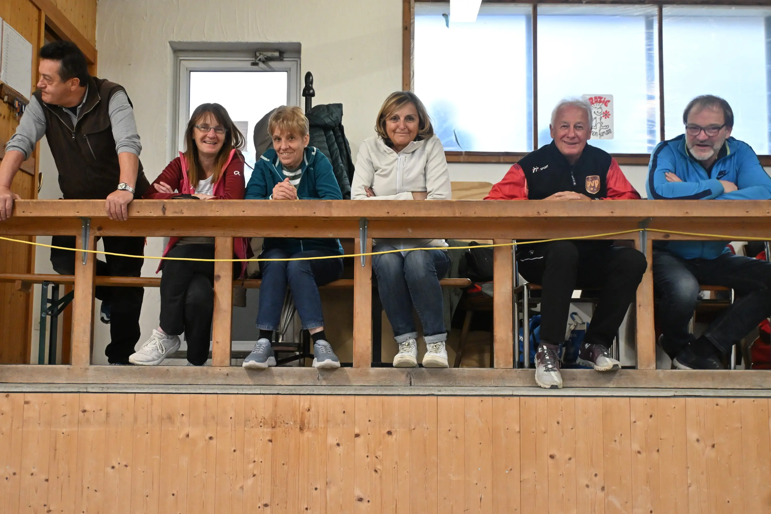 Un gruppo di sei persone sedute su una balaustra, sorridenti e in abbigliamento casual. Alcuni tengono una tazza in mano. Sullo sfondo, ampie finestre che lasciano filtrare la luce.