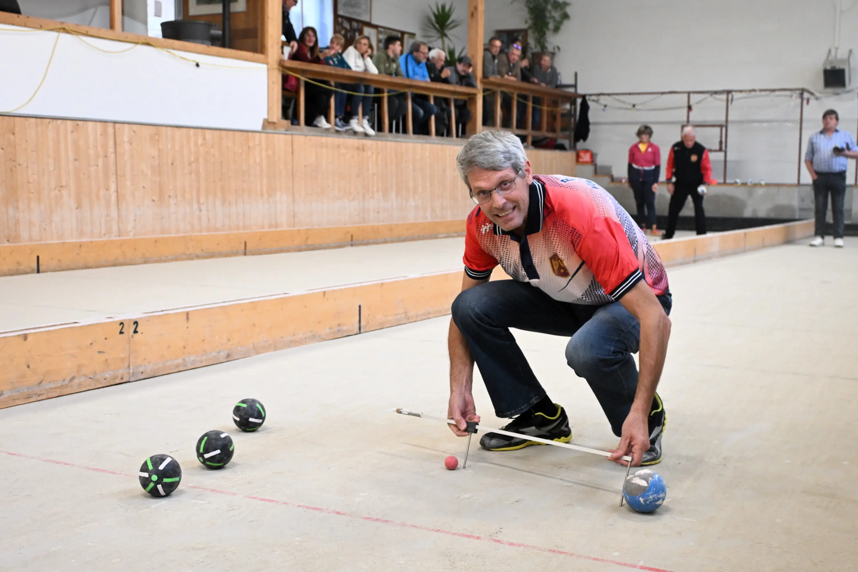 Un uomo piegato in avanti, con occhiali e una maglietta colorata, misura la distanza di una pallina rossa su un campo di bocce. Sul campo ci sono altre palline di diversi colori, mentre in fondo si vedono spettatori che osservano.