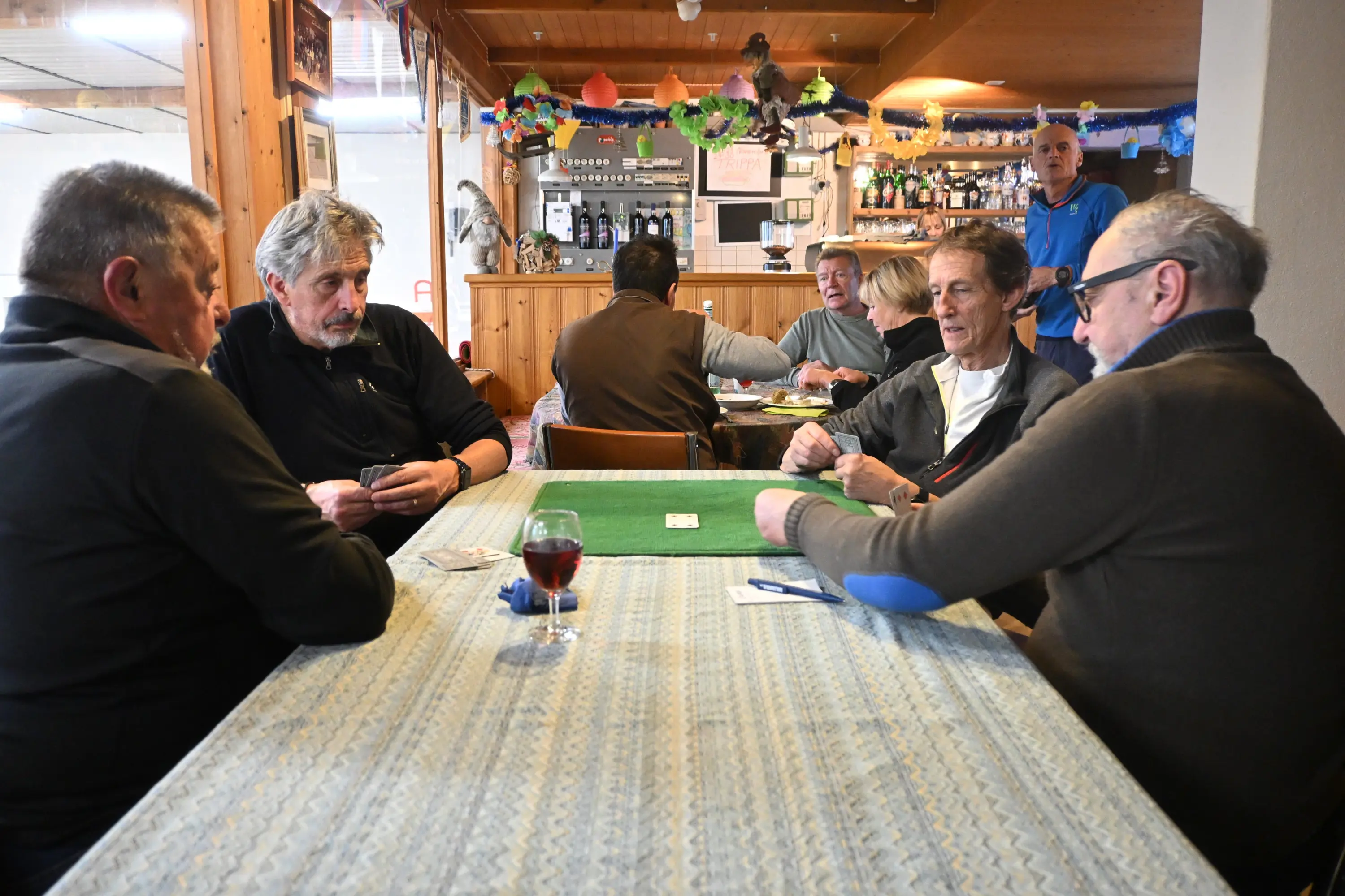 Un gruppo di uomini seduti attorno a un tavolo, mentre giocano a carte. Alcuni clienti sono visibili sullo sfondo, con decorazioni festive. Una bevanda rossa è sul tavolo.