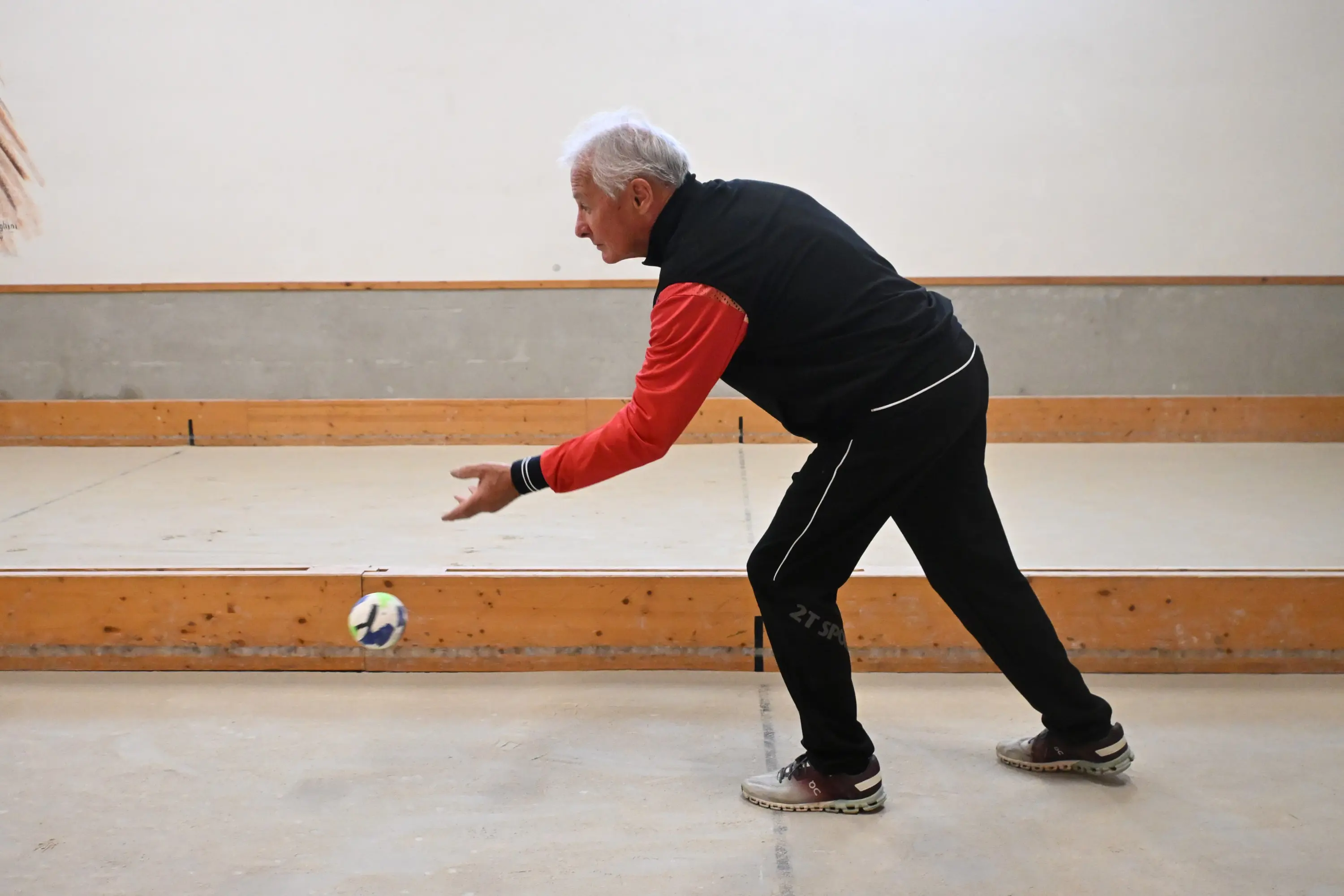 Un uomo anziano sta lanciando una palla in una sala di gioco, vestito con una giacca nera e rossa e pantaloni sportivi neri.