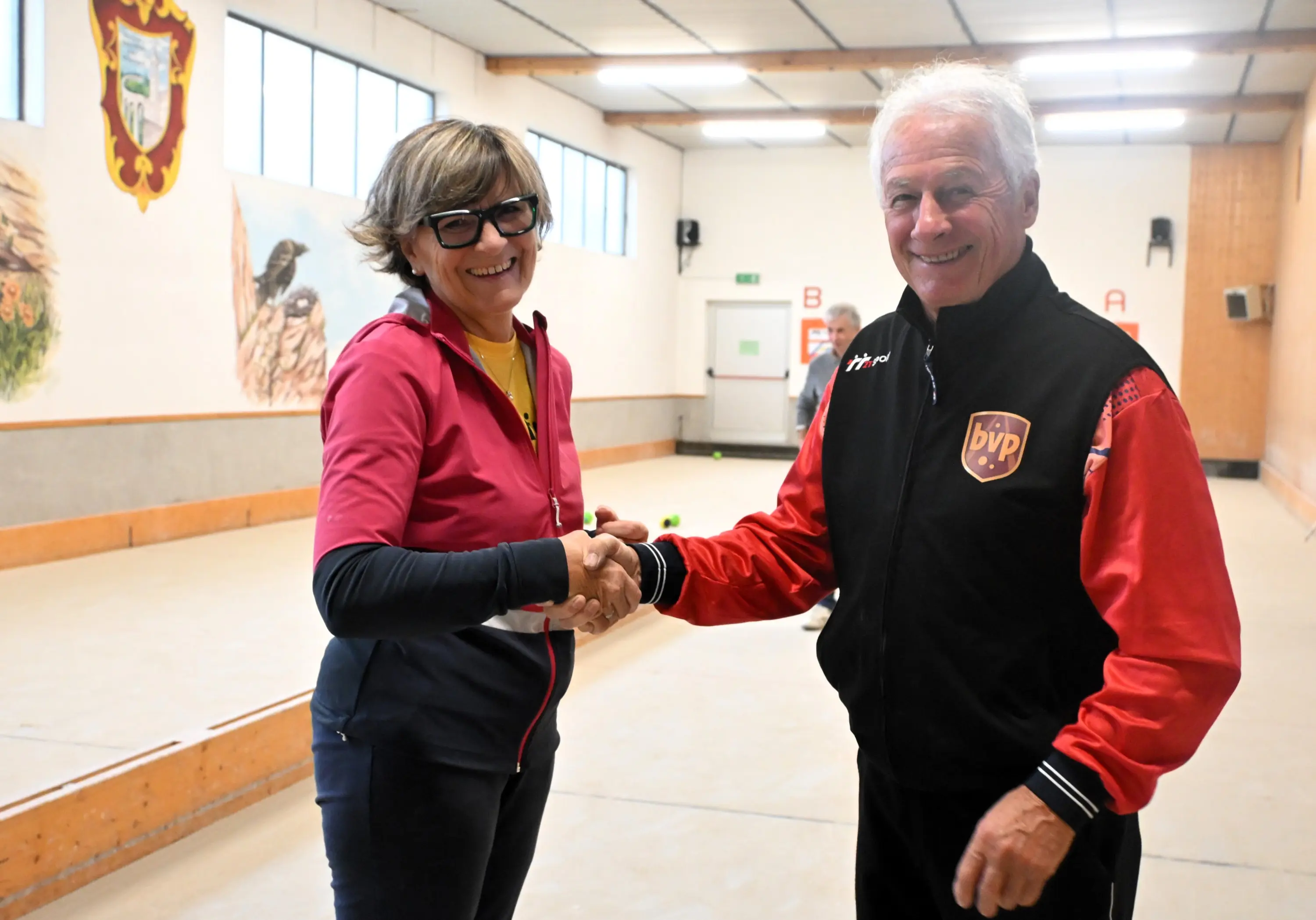 Due persone si stringono la mano in un campo da giochi, sorridendo. Una donna indossa una maglietta gialla sotto una giacca rossa e nera, mentre l'uomo ha una giacca nera con dettagli rossi. Sullo sfondo, si vedono