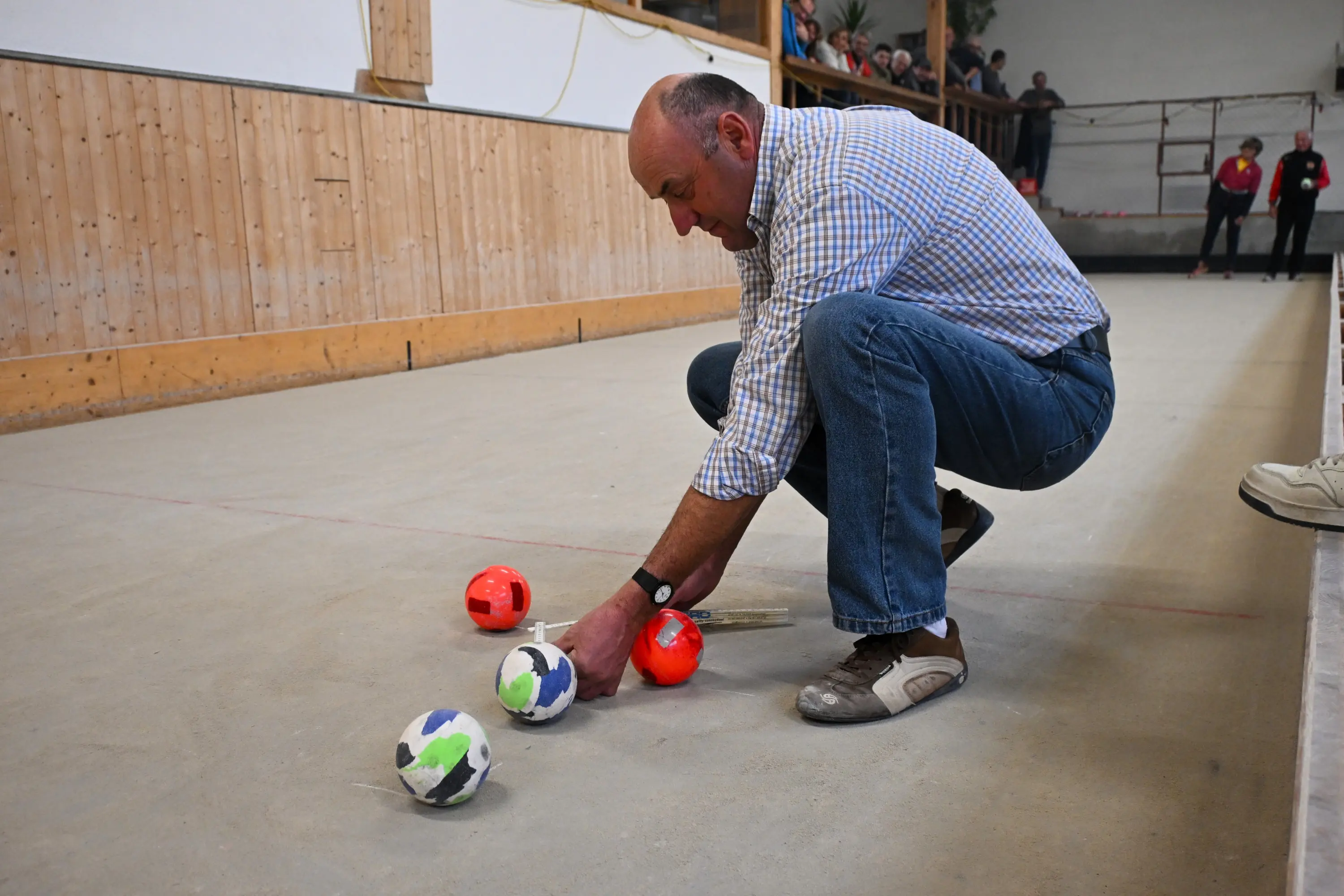 Un uomo si china per sistemare delle sfere colorate su una pista da bocce, mentre altre persone osservano in background.