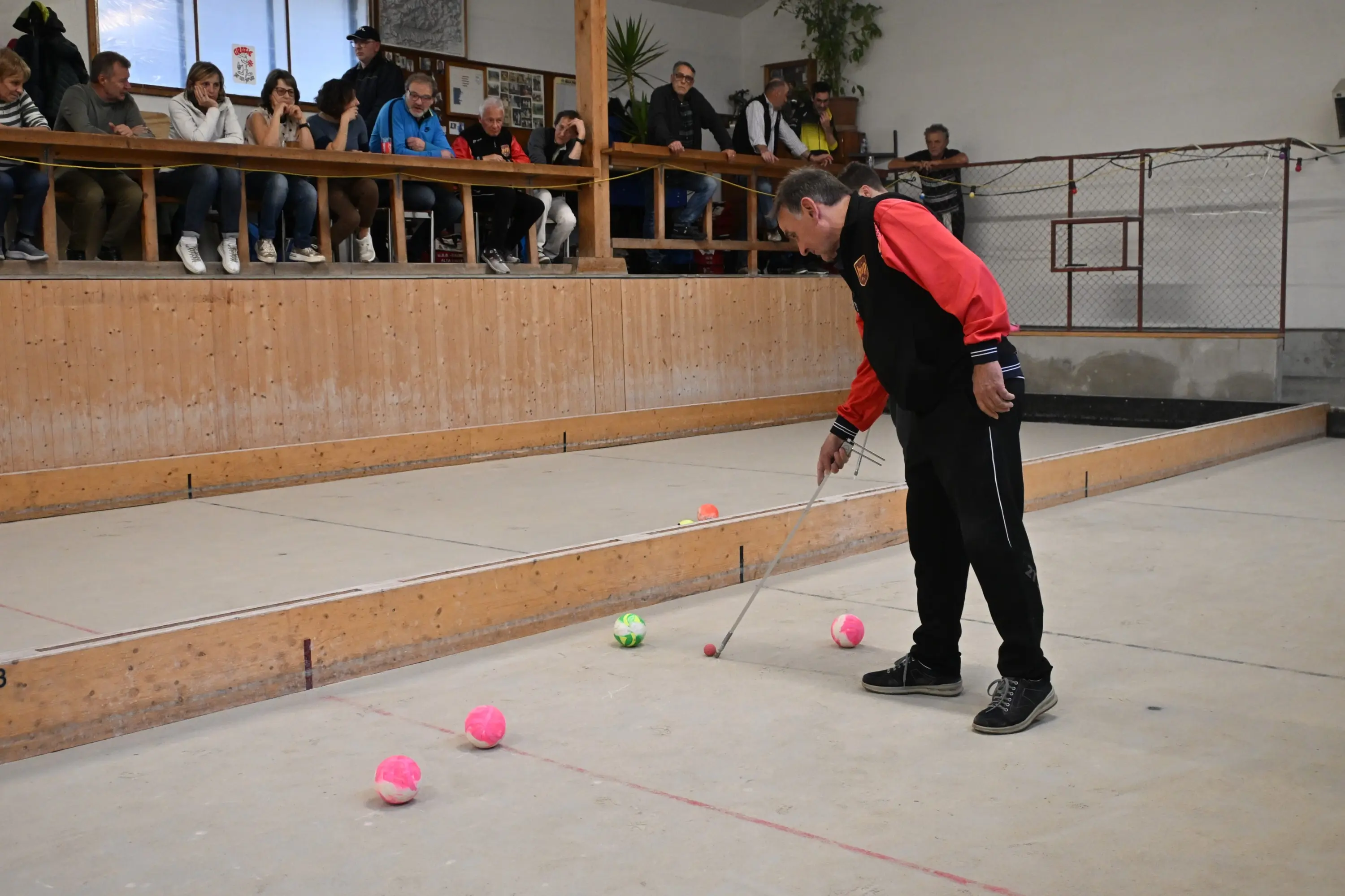 Un uomo adulto, vestito con una giacca nera e rossa, si piega per controllare le palle su una pista da bocce. Sullo sfondo, un gruppo di spettatori osserva attentamente la scena. La pista è in legno e le palle di