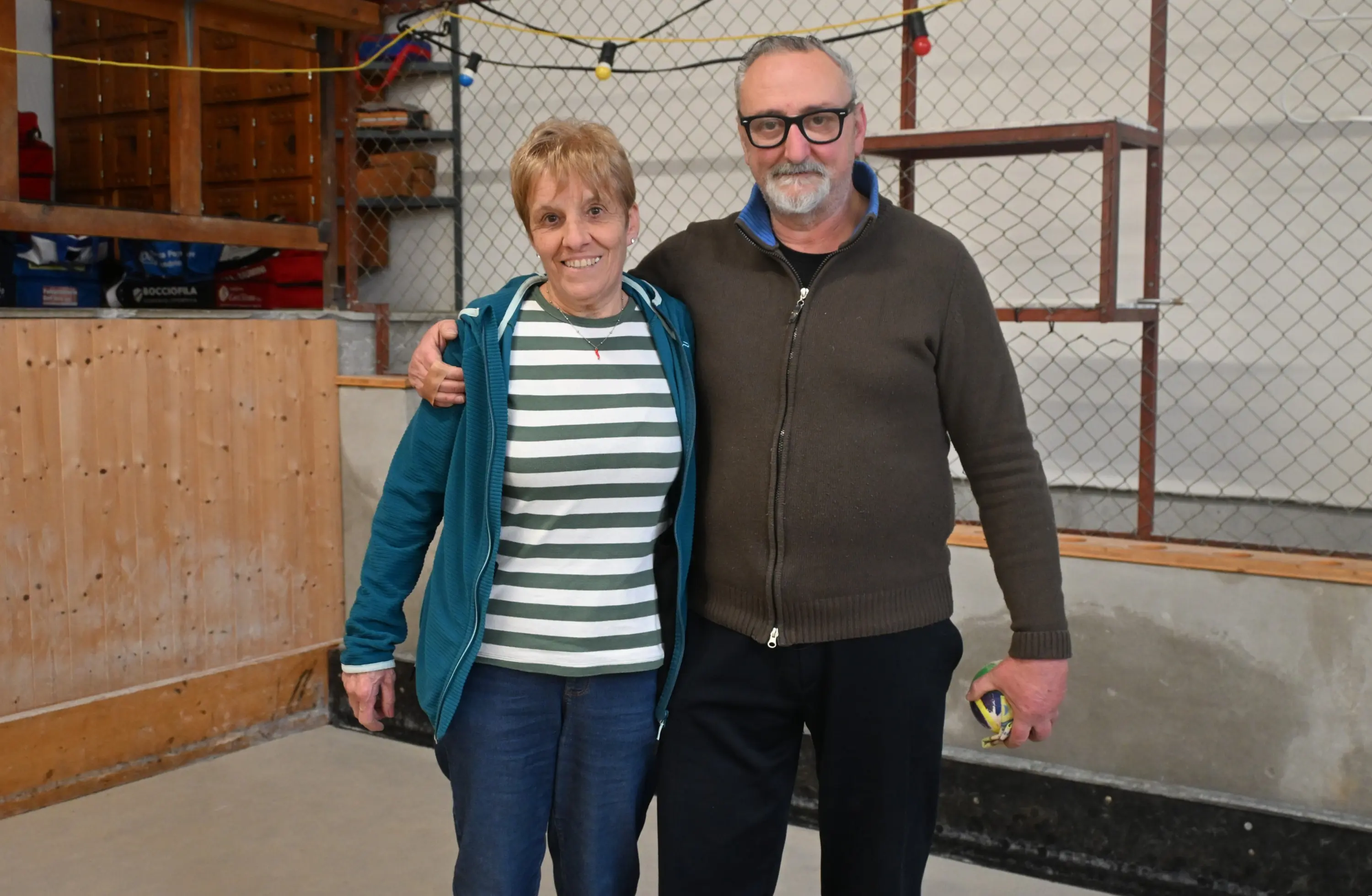 Due persone sorridenti si abbracciano in un ambiente interno con pareti di legno e rete. Una donna indossa una maglietta a righe e una giacca blu, mentre l'uomo ha un maglione grigio e occhiali.