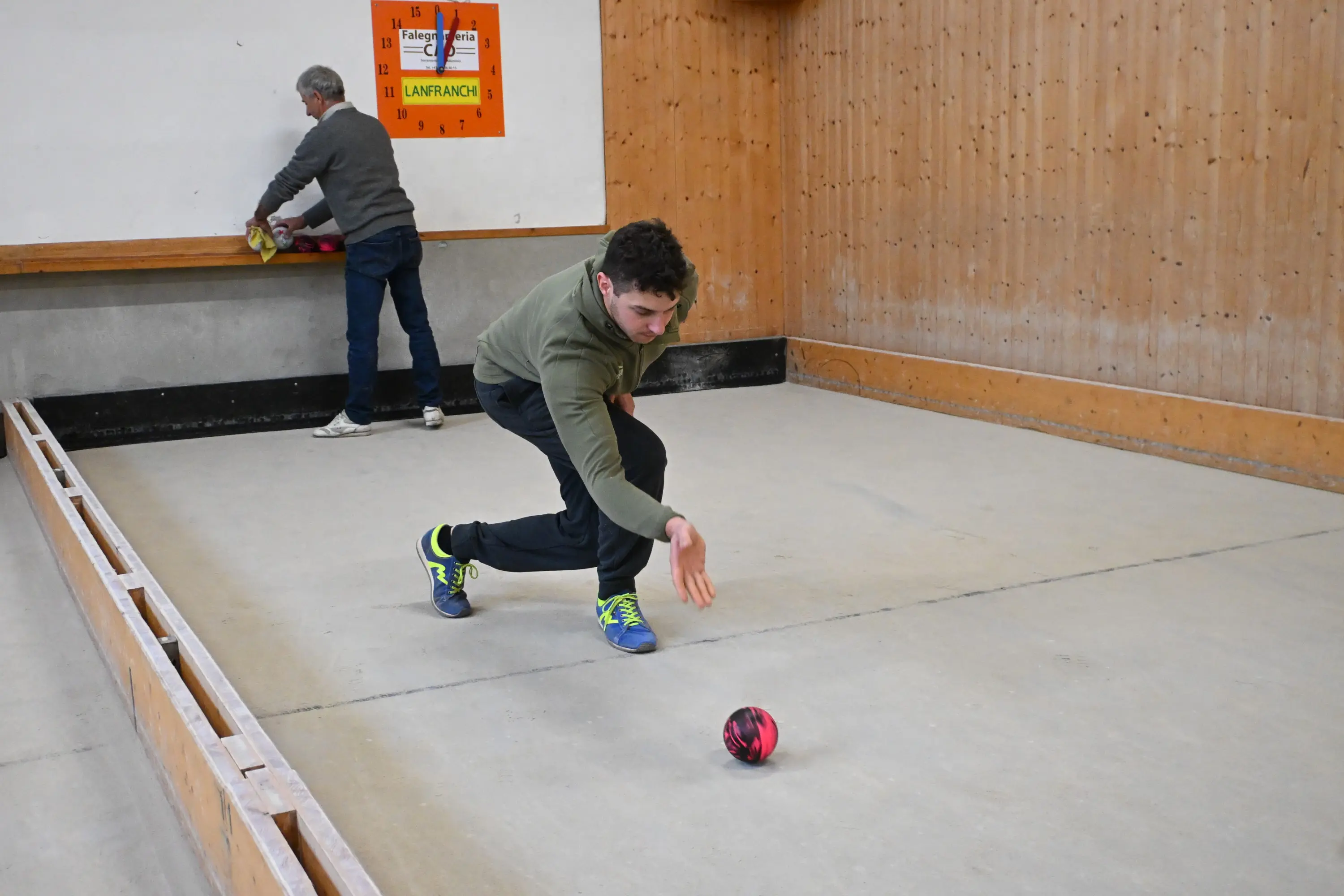 Un uomo sta lanciando una boccia su un campo di bocce, mentre un altro uomo si trova sullo sfondo, intento a preparare il suo tiro.