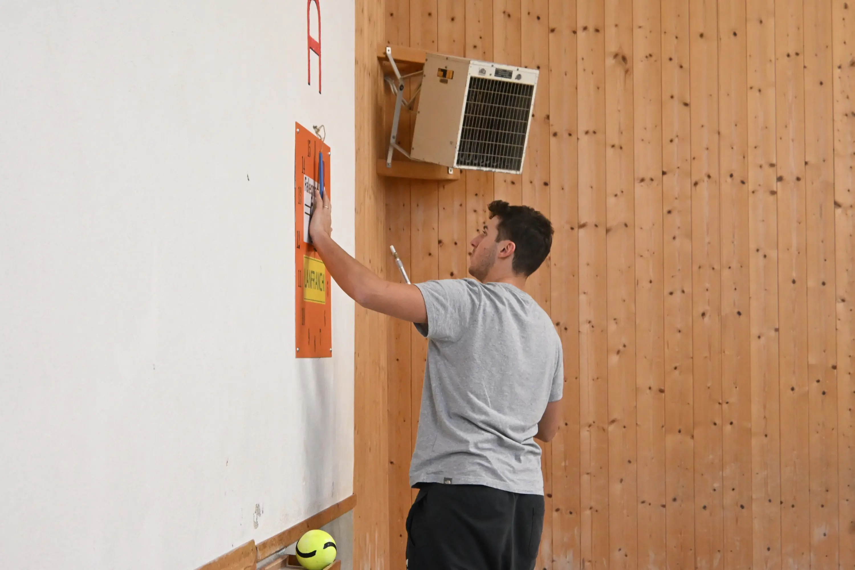 Un giovane sta dipingendo un muro in una palestra, utilizzando un rullo per applicare colore. Sul pavimento, un pallone da pallavolo è parzialmente visibile. La parete presenta un rivestimento in legno e un'unità di aria condizion
