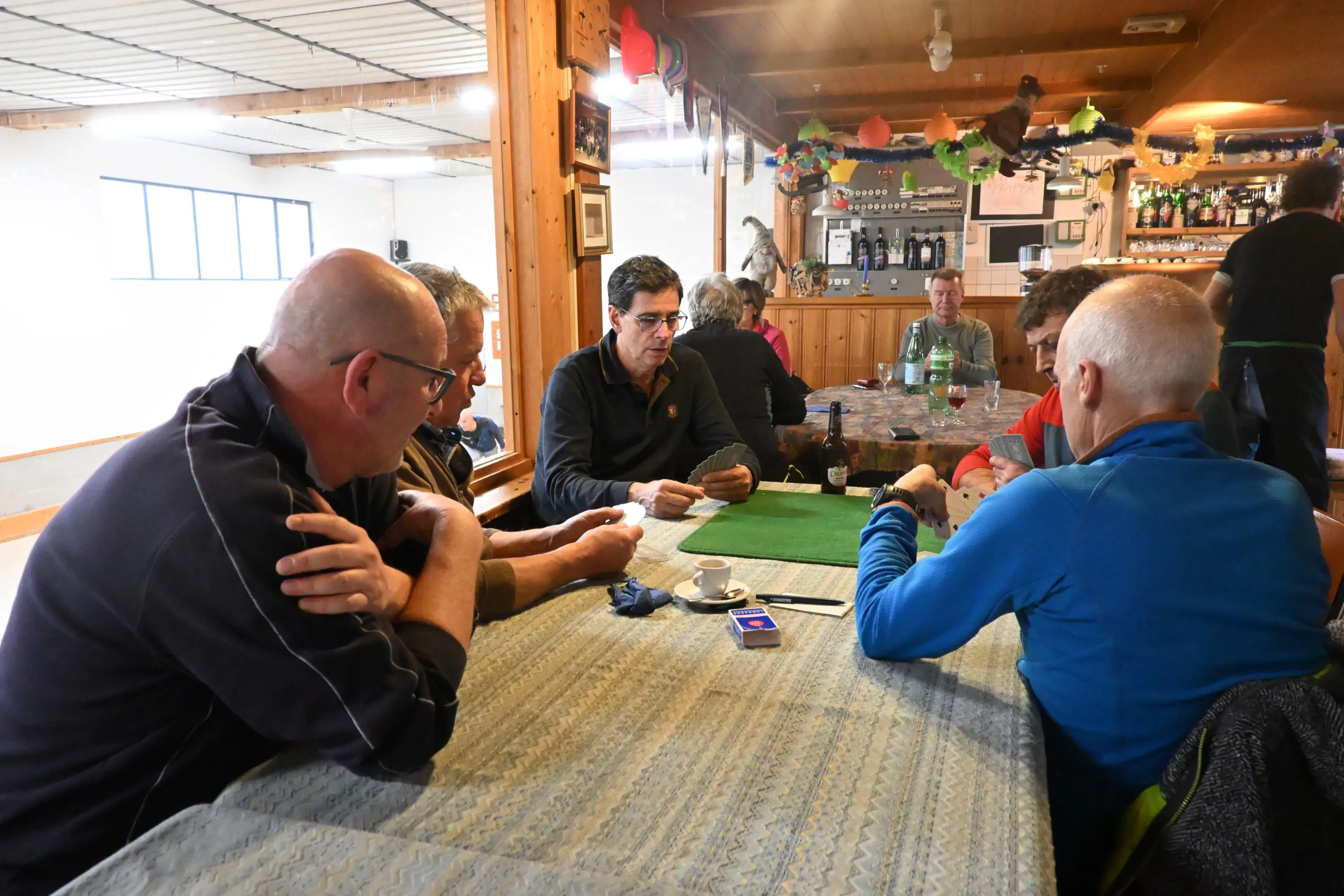 Un gruppo di uomini seduti attorno a un tavolo, impegnati in un gioco di carte in un ambiente conviviale. Sullo sfondo si vedono altre persone che socializzano in un locale ben arredato.