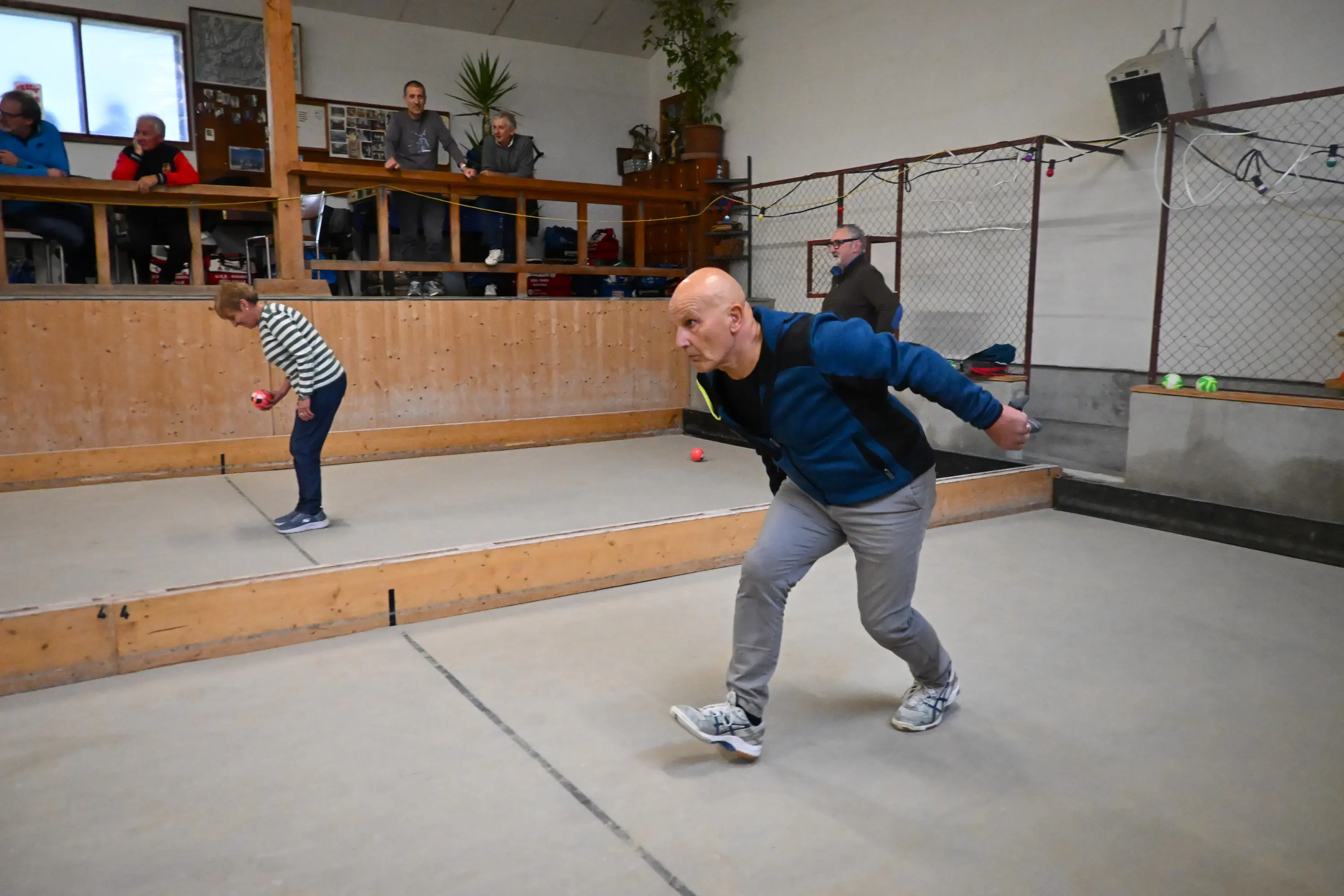 Un uomo lancia una palla in una sala da bowling mentre un altro osserva. Sullo sfondo, alcune persone seguono il gioco da un’idea rialzata. Il pavimento è in cemento e ci sono piante ornamentali nella stanza.