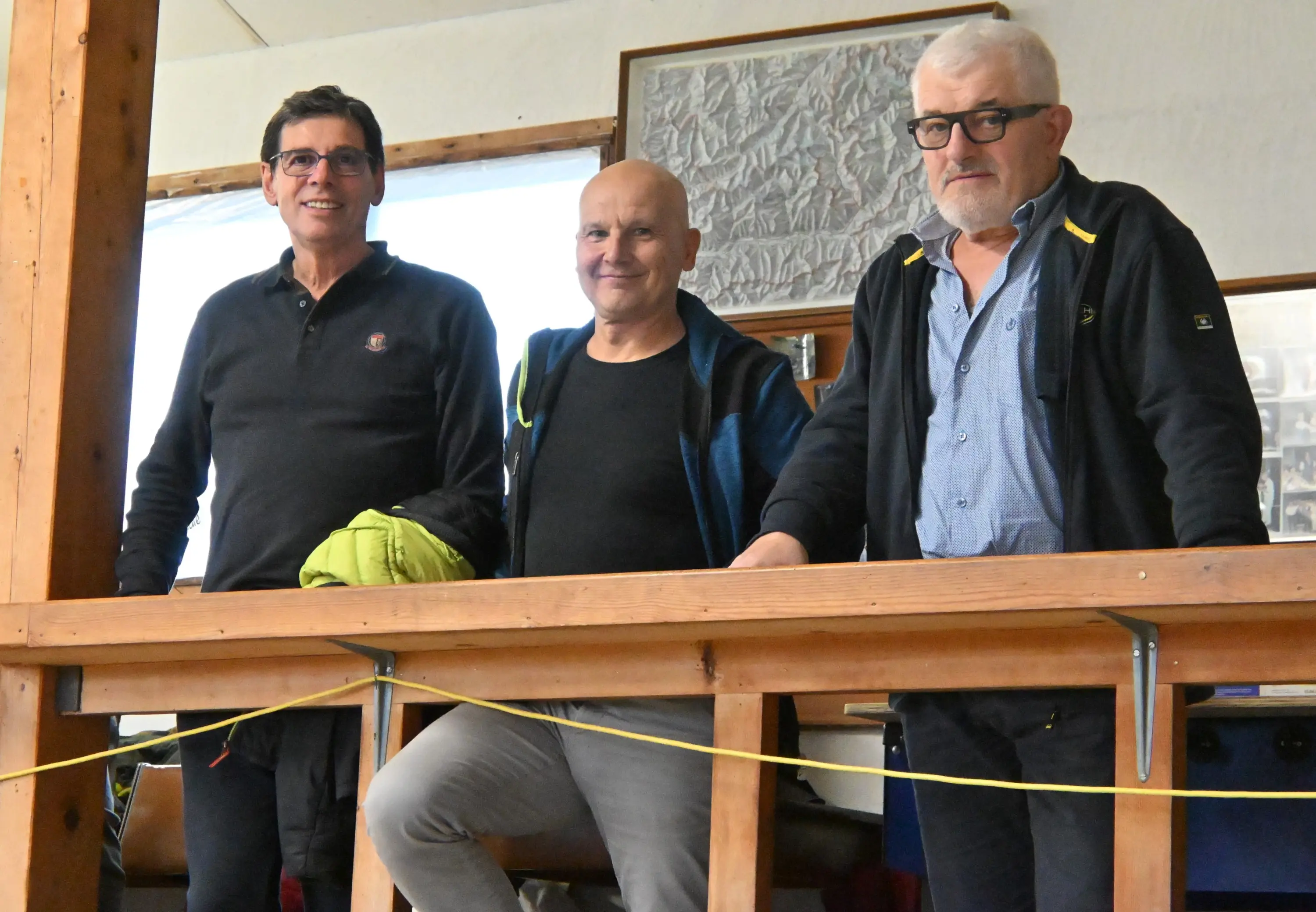 Tre uomini sorridenti in un ambiente interno, appoggiati su un parapetto in legno. Due indossano occhiali e uno ha una giacca nera. Sullo sfondo, si intravedono elementi di legno e una parete decorata.