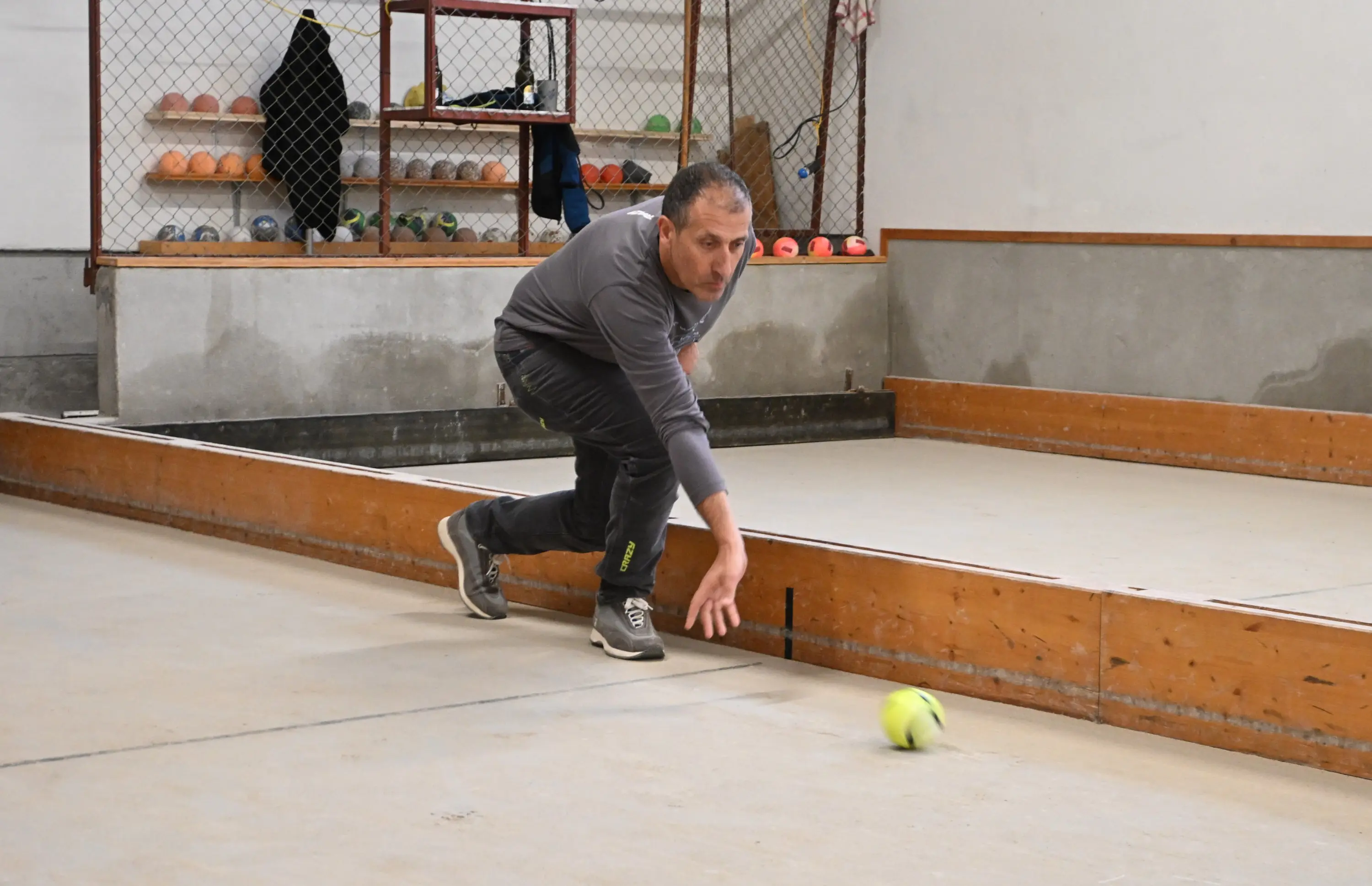 Un uomo si china per colpire una pallina da bocce su un campo di bocce. Indossa abbigliamento sportivo e sembra concentrato mentre lancerà la pallina. Sullo sfondo si vedono palline e attrezzi sistemati su una piattaforma.