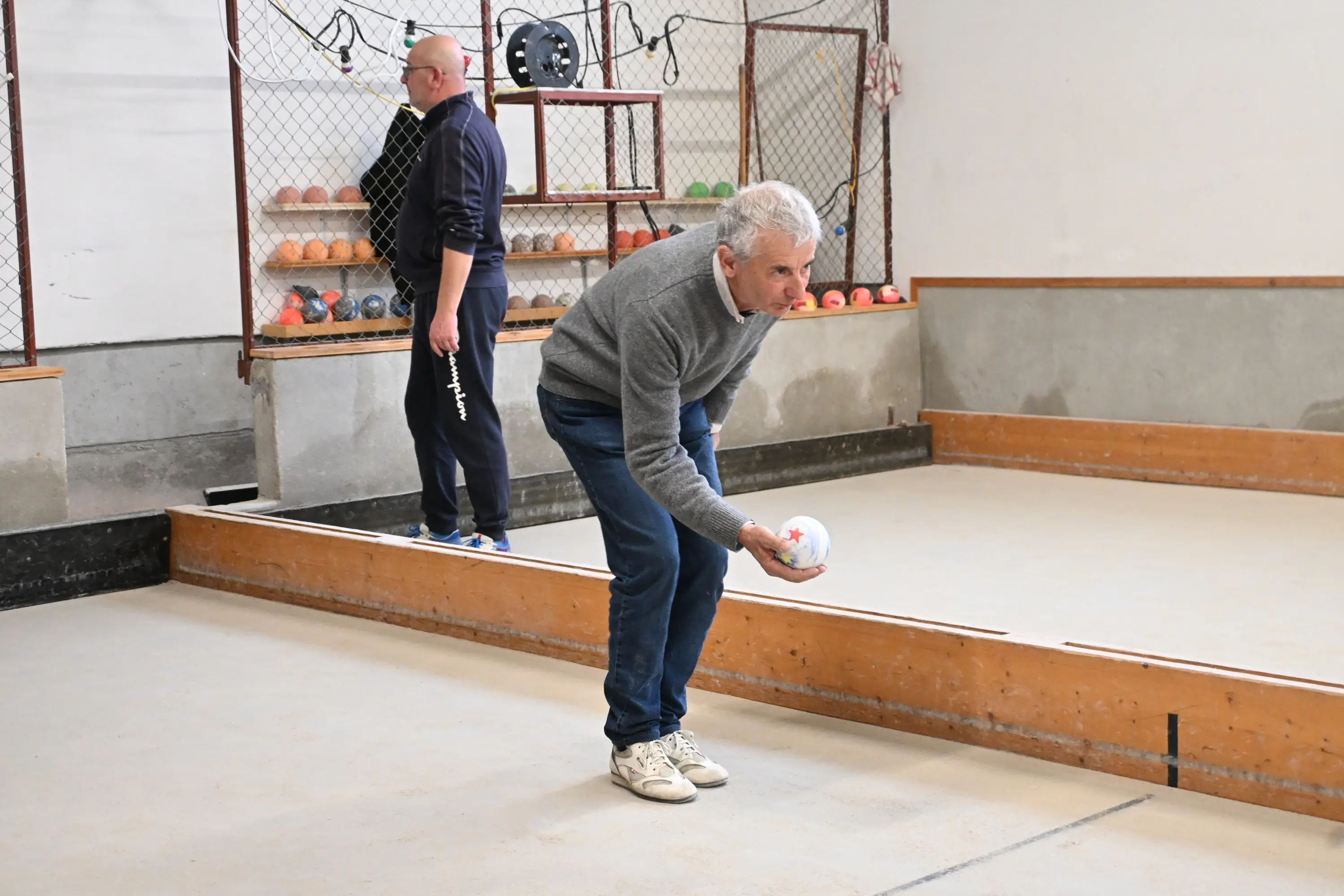 Due uomini in un campo di bocce. Uno, con i capelli bianchi, si piega in avanti mentre lancia una boccia. L'altro, vestito di nero, osserva in secondo piano. Sullo sfondo si vedono altre bocce colorate.