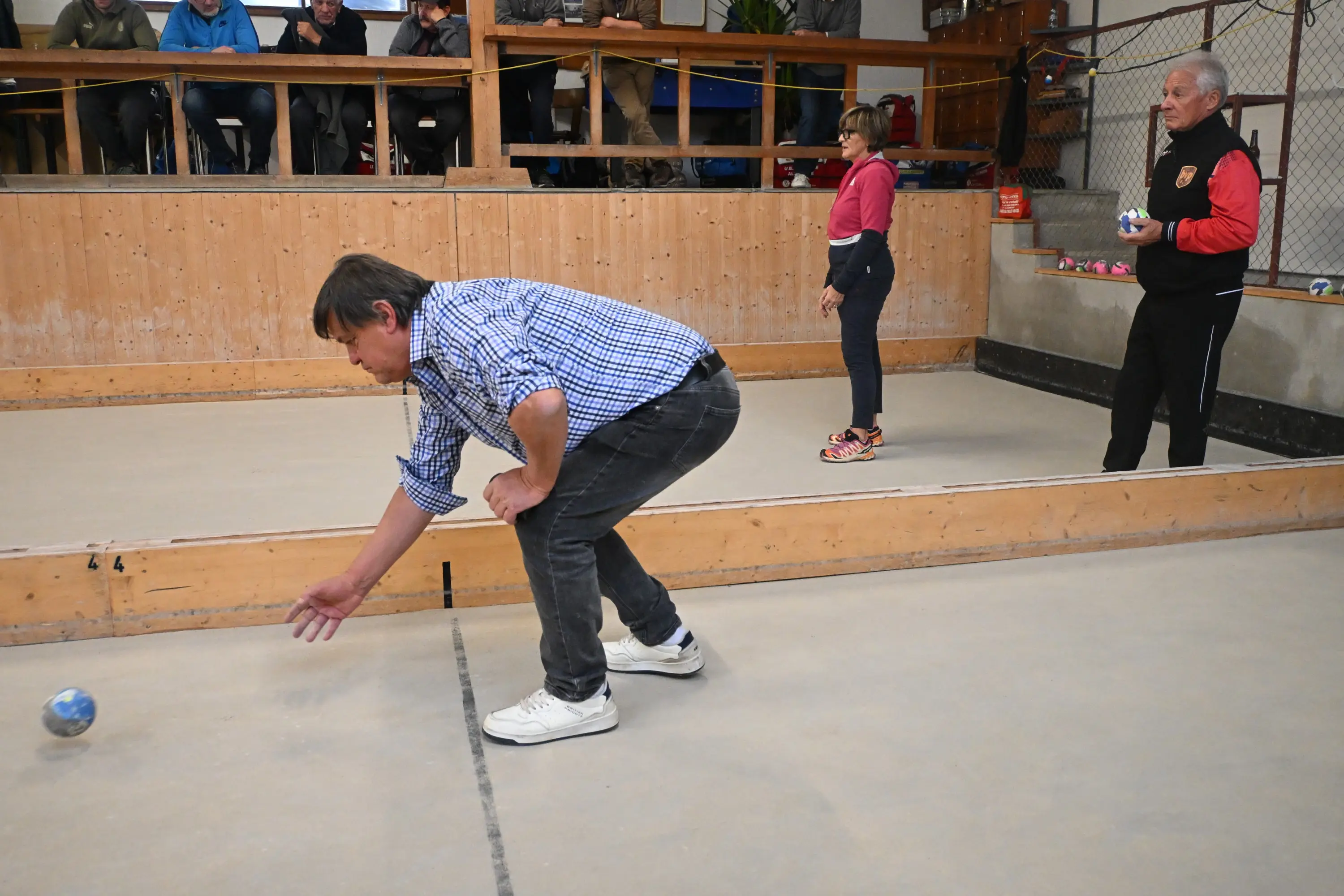 Giocatori di bocce in un campo coperto, con un uomo chino che lancia una boccia verso la linea di partenza. Una donna in piedi osserva, mentre un'altra persona è visibile sullo sfondo. Il pubblico è seduto in tribuna.