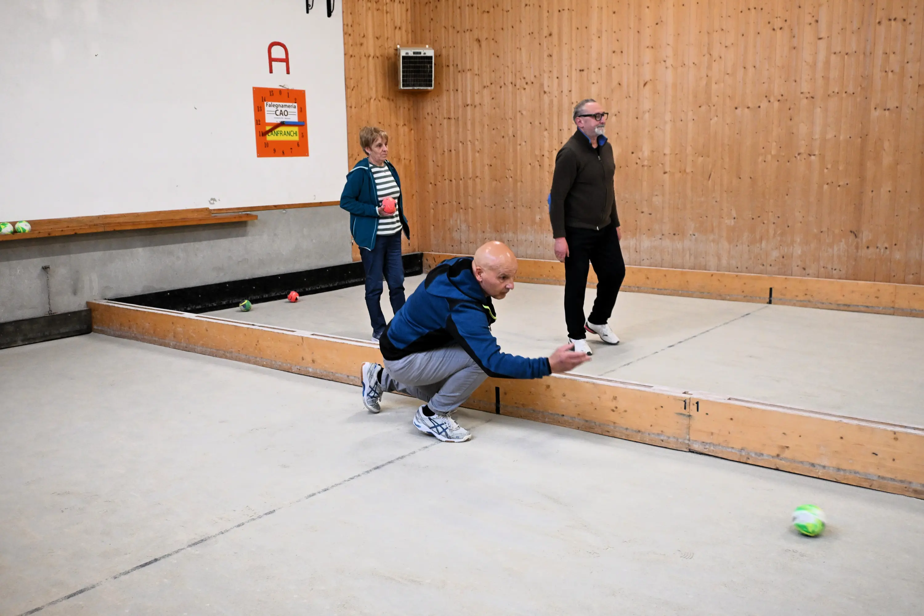 Un uomo lancia una boccia mentre due persone osservano in un campo da bocce. Il pavimento è in cemento e le pareti sono in legno. Si notano alcune bocce colorate sparse sul terreno.