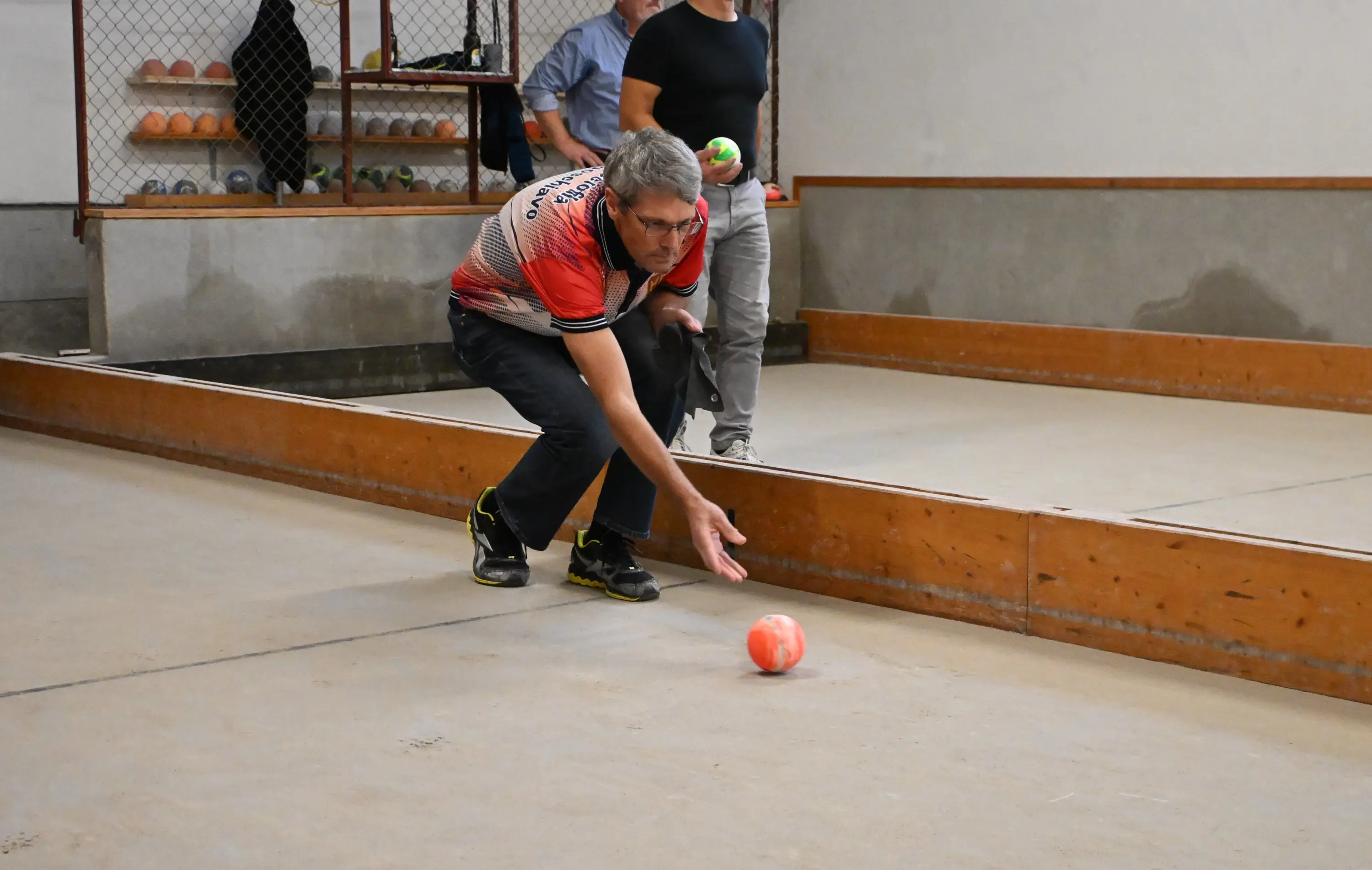 Un uomo gioca a bocce, mentre si inginocchia per lanciare la pallina verso il bersaglio. Sullo sfondo, altre persone osservano e partecipano al gioco.