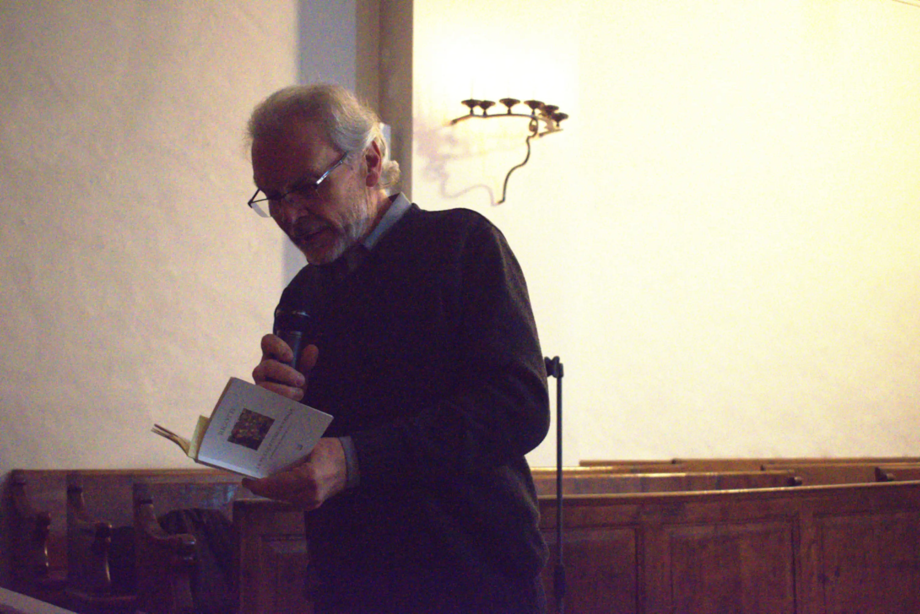 Uomo anziano con occhiali, vestito con un maglione scuro, legge da un libro mentre tiene un microfono in mano. Sullo sfondo, un ambiente interno semplice