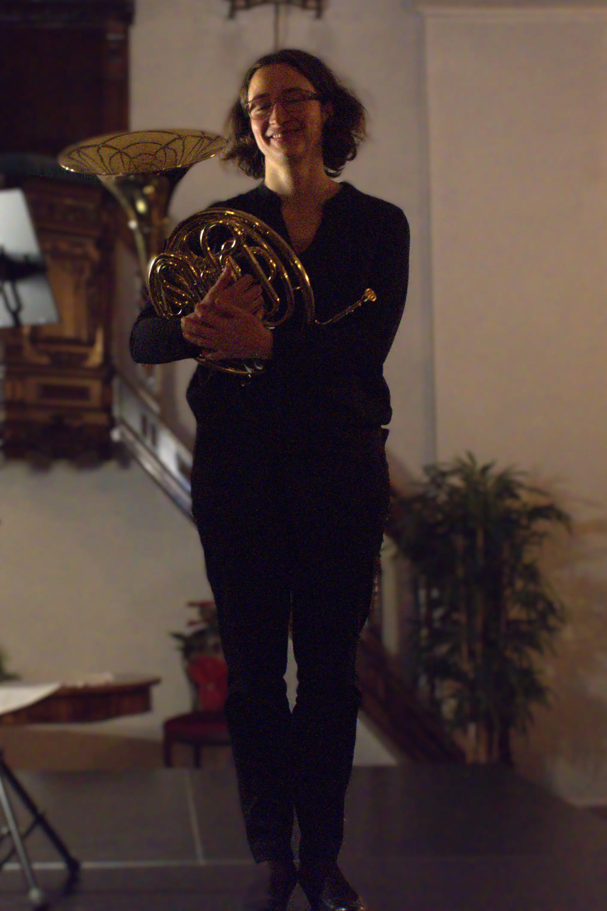 Una musicista sorride mentre tiene in braccio un corno francese. Indossa un abbigliamento scuro e si trova su un palcoscenico, con decorazioni sullo