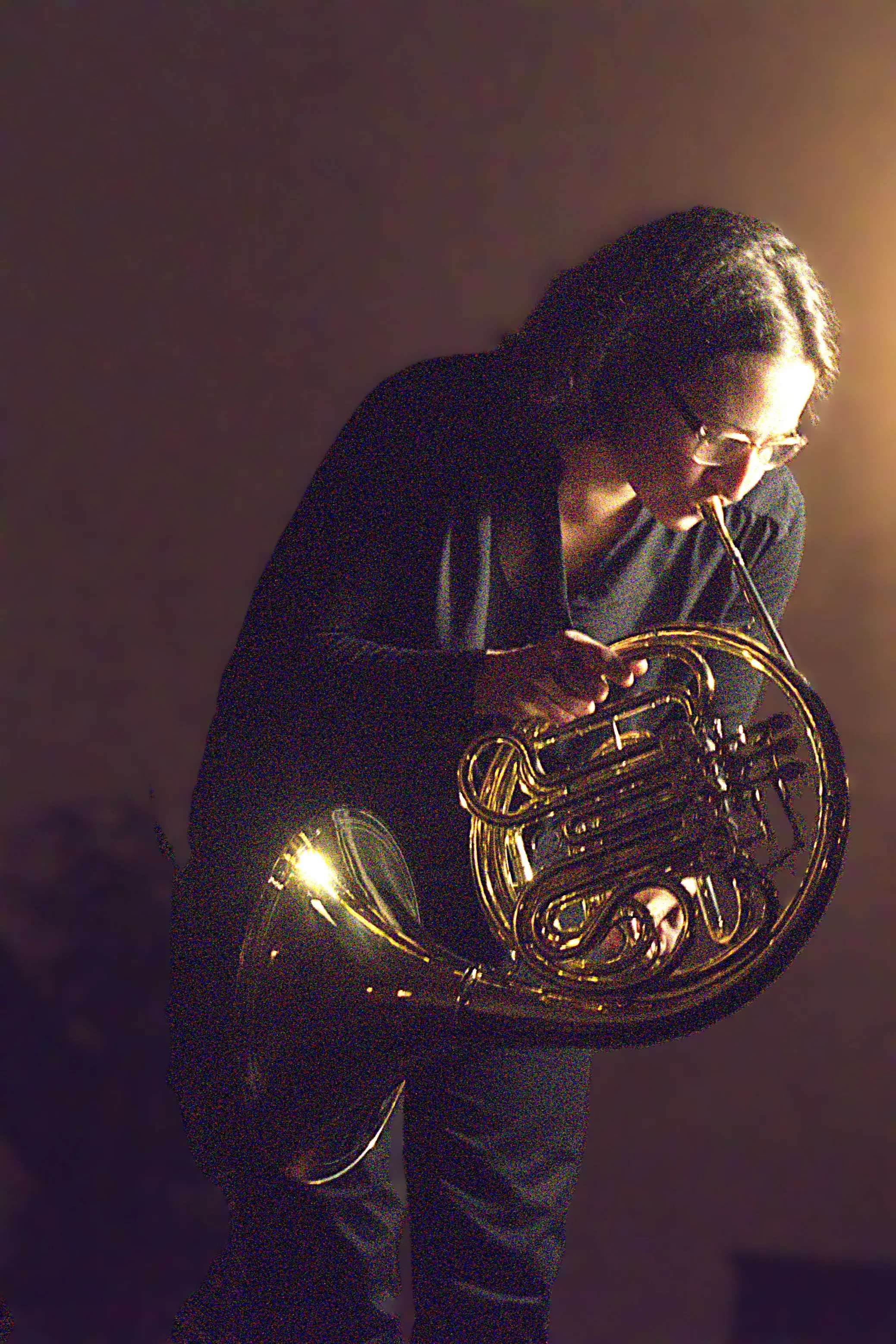 Una musicista suona il corno francese, piegata in avanti, con una luce calda che illumina l'ottone dellstrumento e riflette sulle sue lenti. Ind