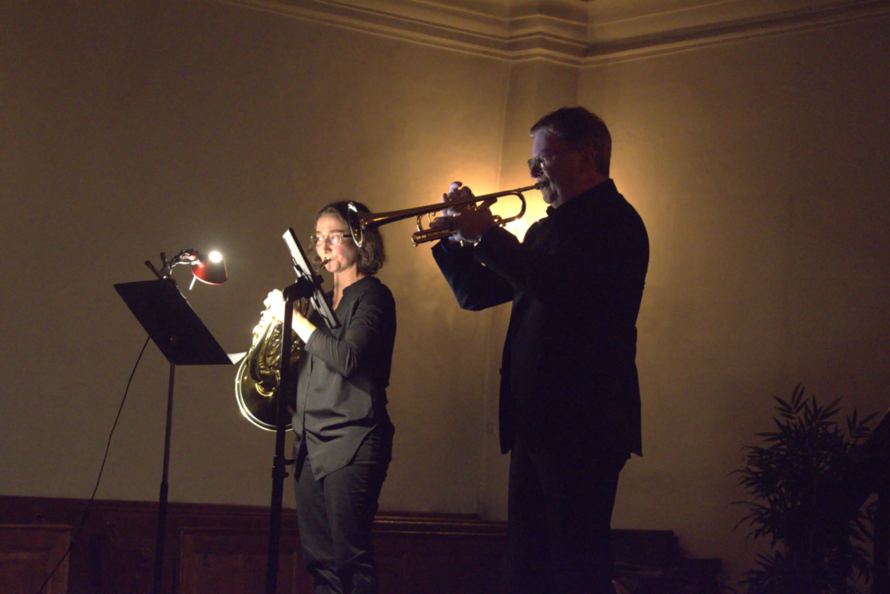 Due musicisti suonano in scena, uno con un trombone e l'altro con una tromba. Sono illuminati da una luce soffusa. Sullo sfondo, si intrav