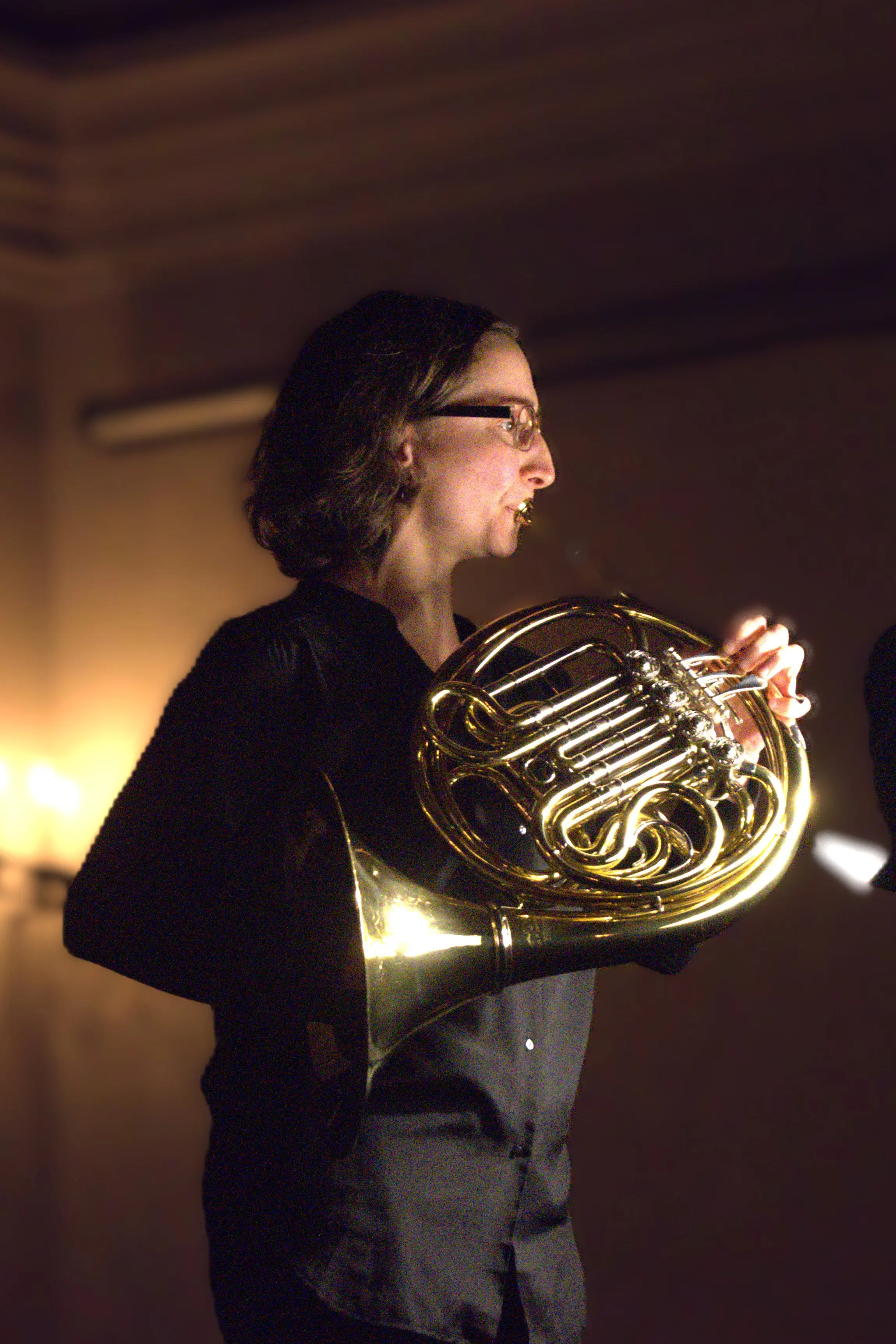Una musicista suona un corno francese, indossando una camicia nera e occhiali. Lo sfondo è sfocato e illuminato da luci calde.