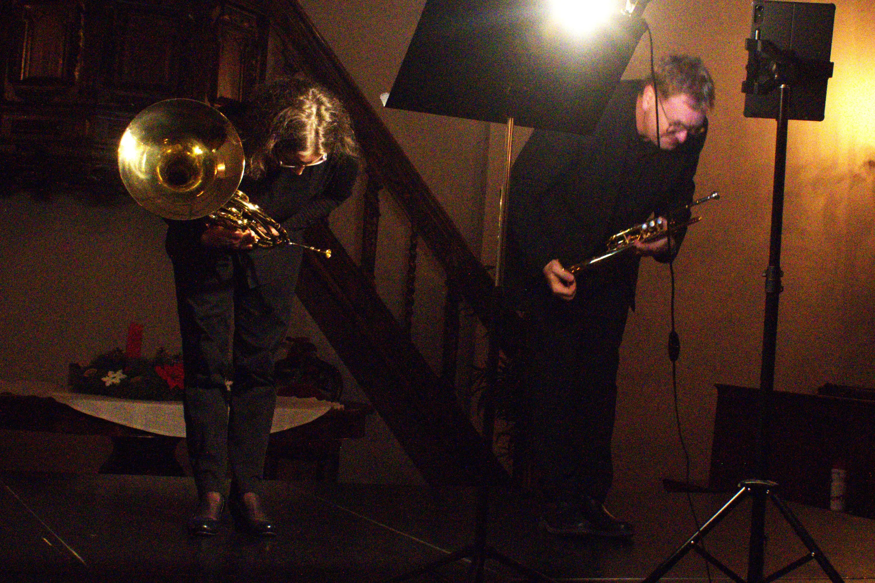 Due musicisti sul palco, piegati in segno di gratitudine, uno con una tromba e l'altro con un trombone. Sullo sfondo, una scala in legno