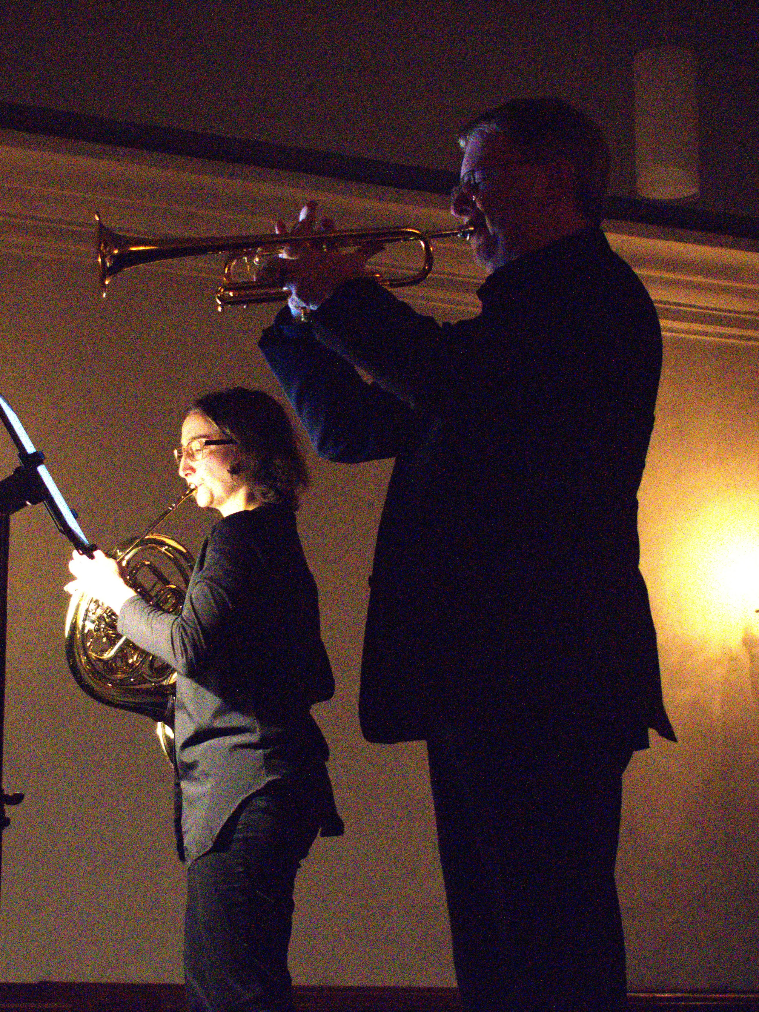 Un musicista suona la tromba mentre una musicista interpreta il corno francese. Entrambi sono in silhouette, con luci soffuse sullo sfondo.