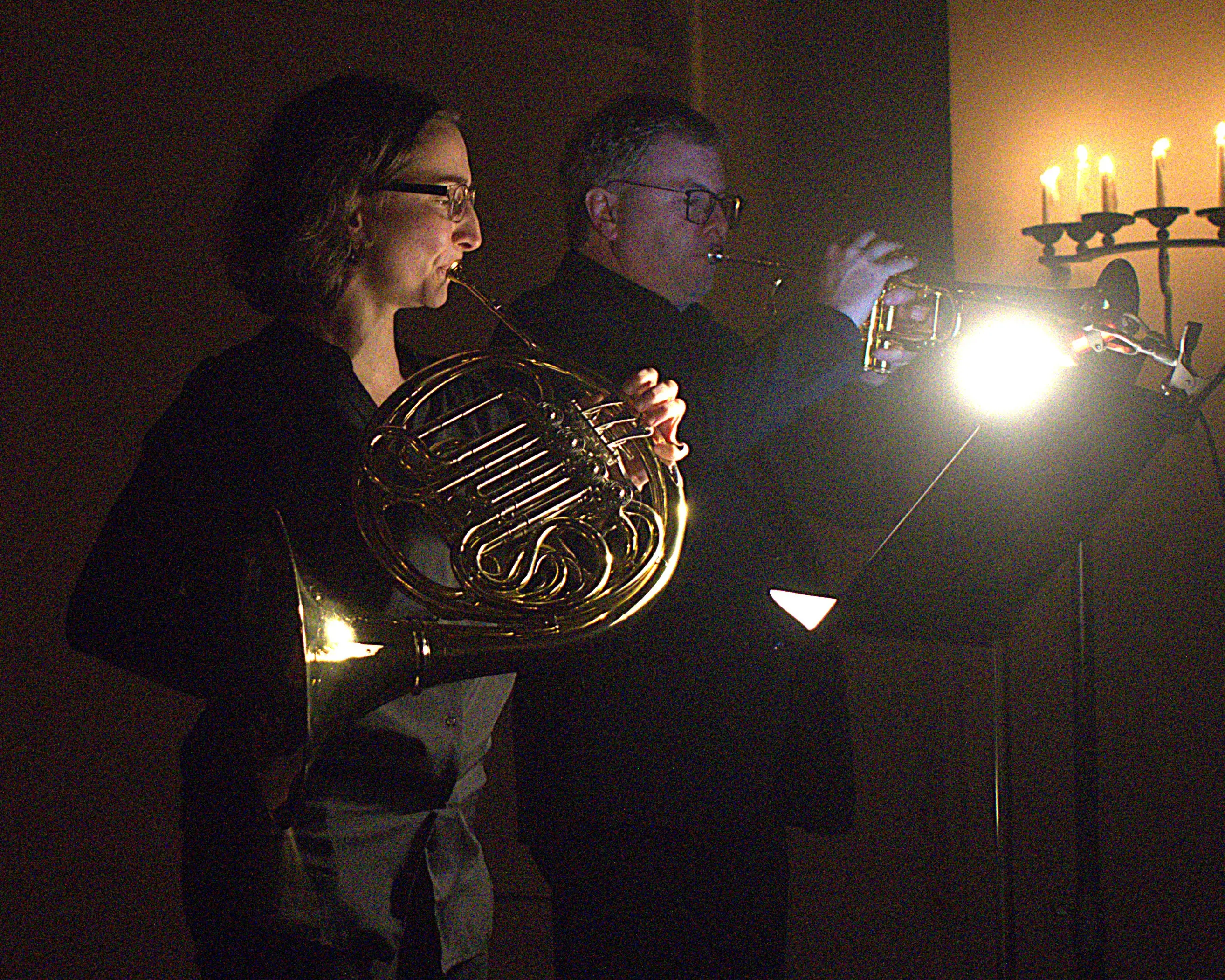 Due musicisti suonano strumenti musicali in un ambiente poco illuminato. Una donna con occhiali tiene un corno francese, mentre un uomo, in secondo piano, suona una