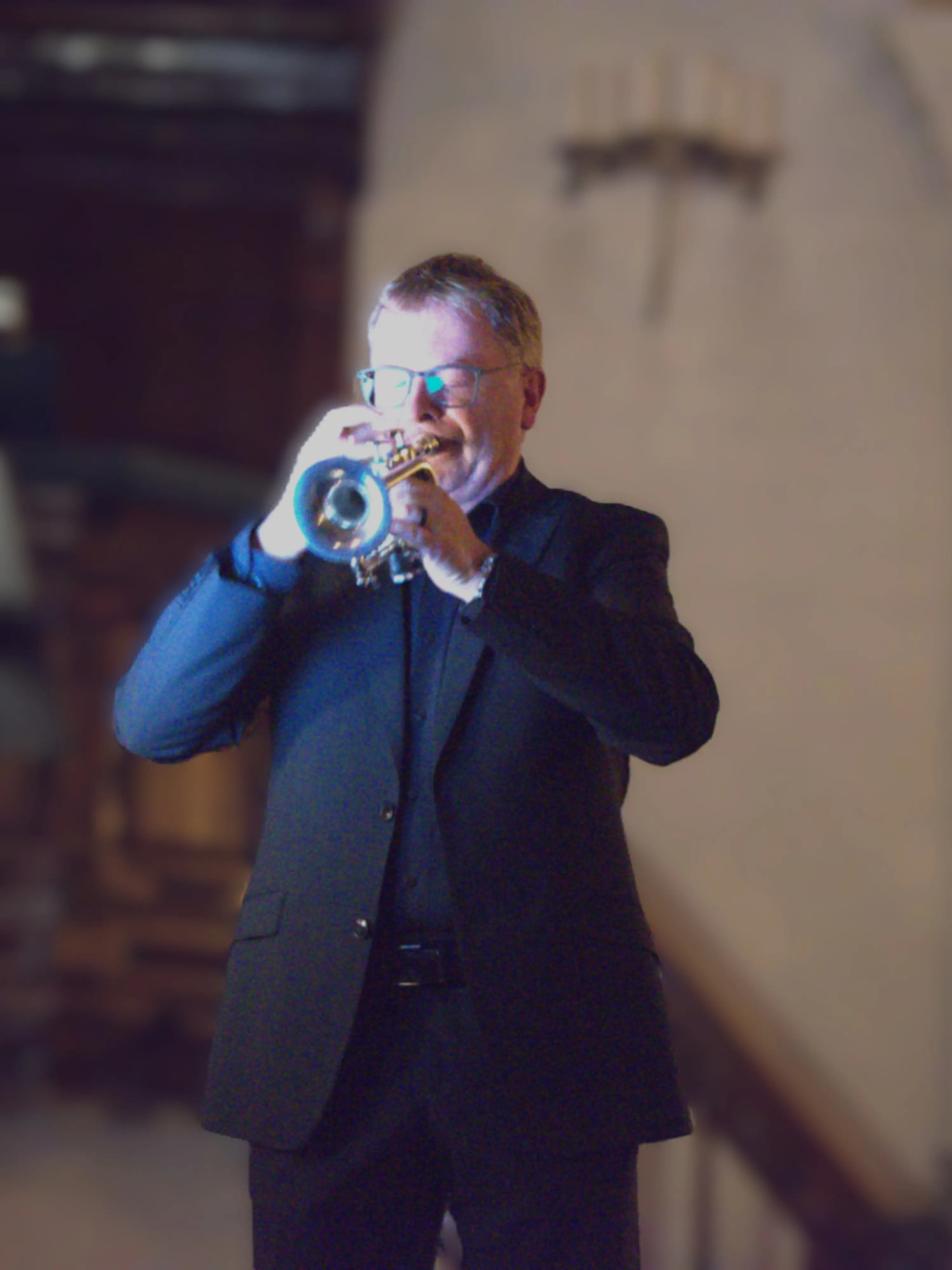 Un musicista in abito nero suona la tromba, sorridendo, in un ambiente elegantemente illuminato.