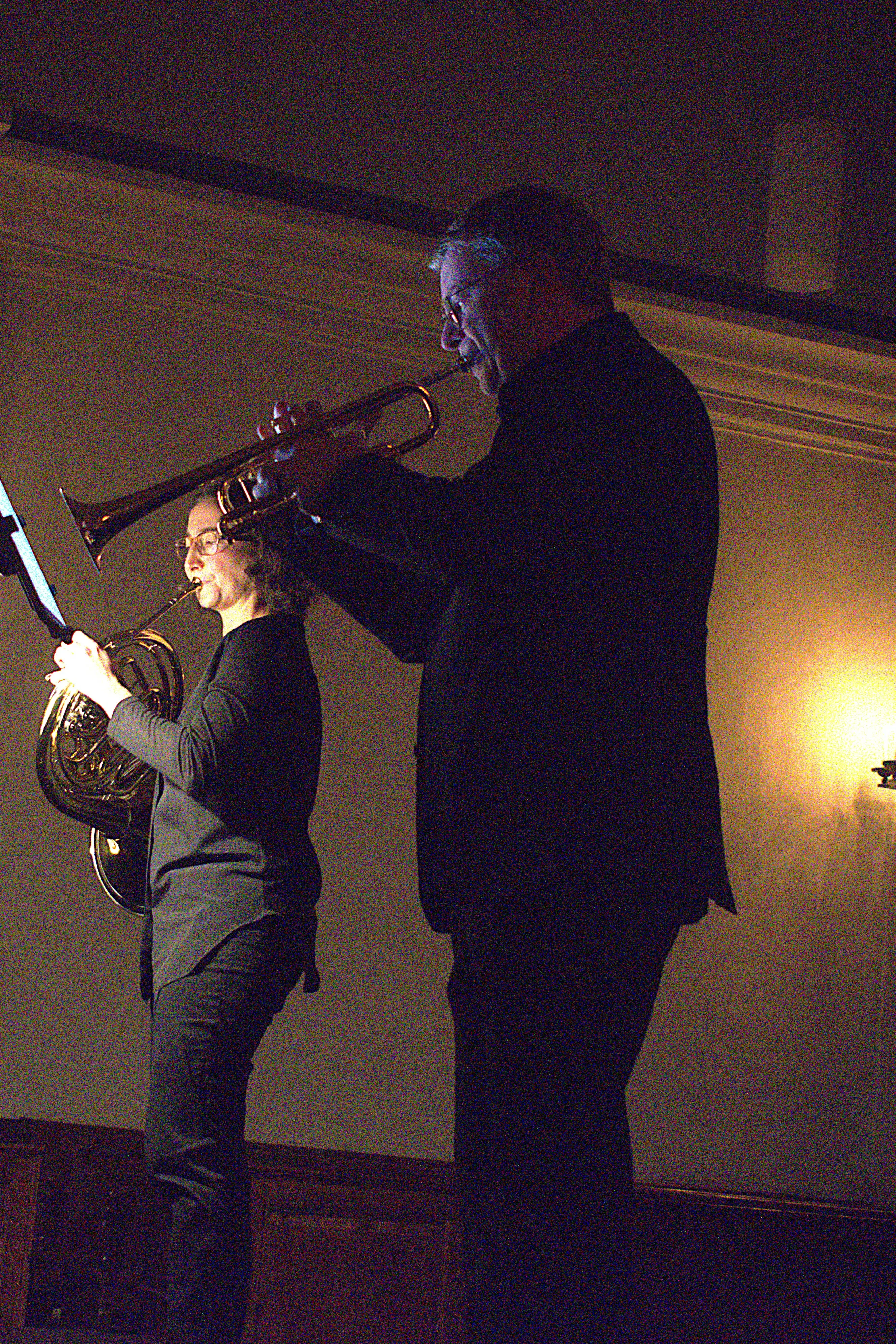 Due musicisti si esibiscono su un palcoscenico. Un uomo suona una tromba, mentre una donna suona un corno francese. La scena è illuminata da luc