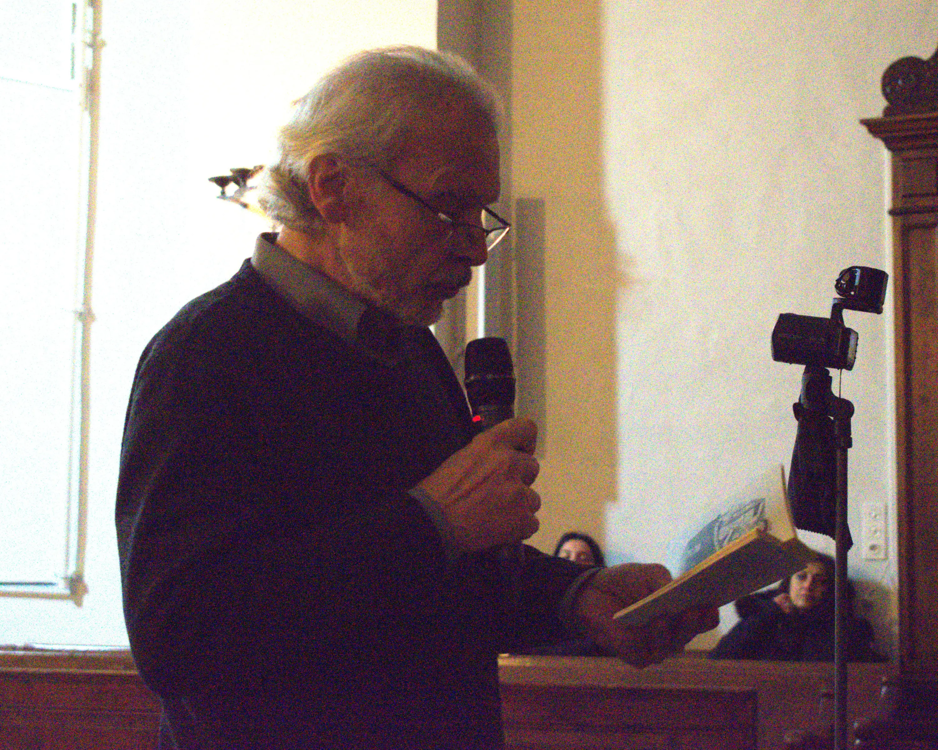 Un uomo anziano legge un libro in una sala, tenendo un microfono in mano. Sullo sfondo, si intravedono volti di persone sedute che lo ascoltano attent