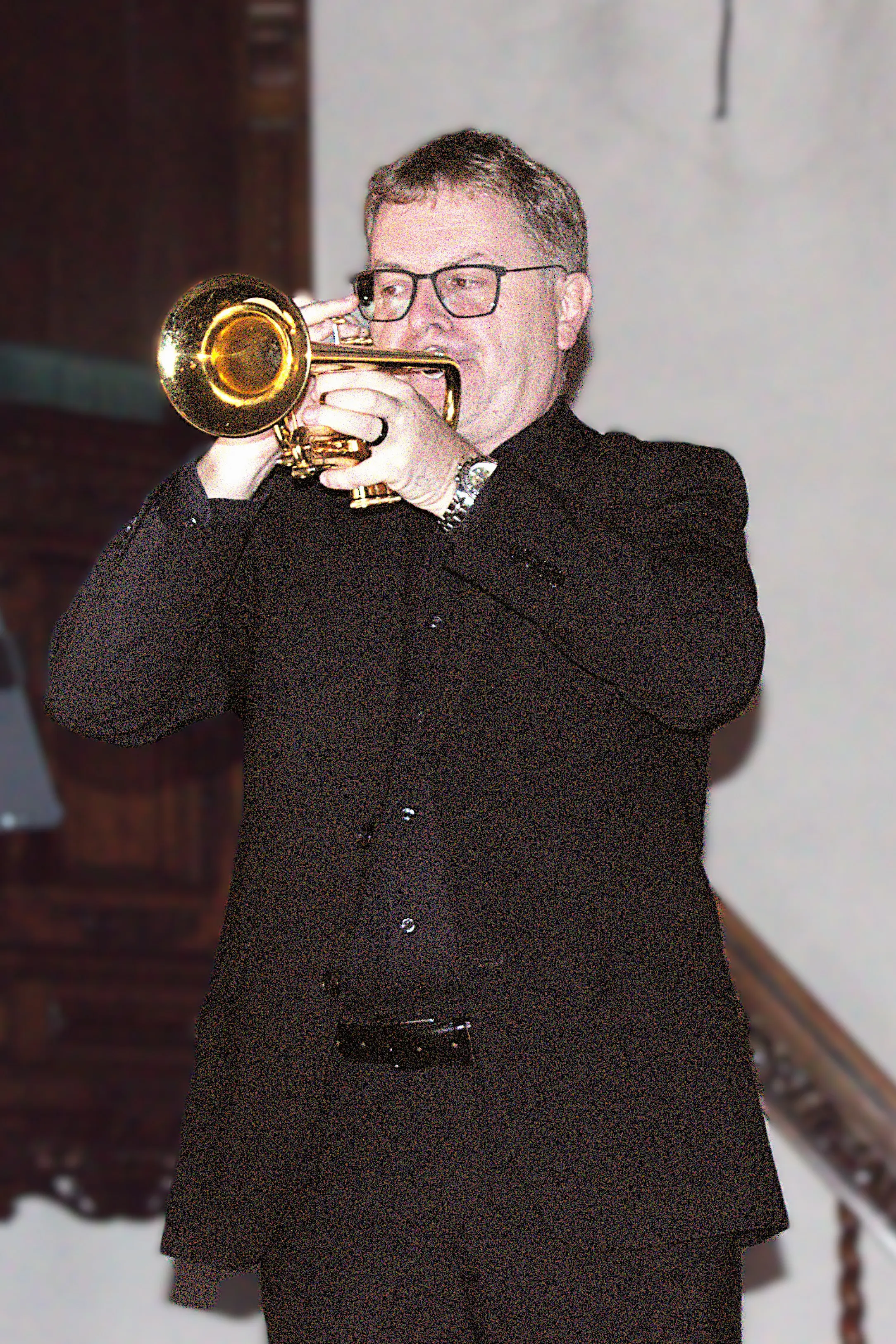 Un uomo in abbigliamento scuro suona una tromba dorata, concentrato e con gli occhiali, mentre si trova su un palco. Sullo sfondo si intrav