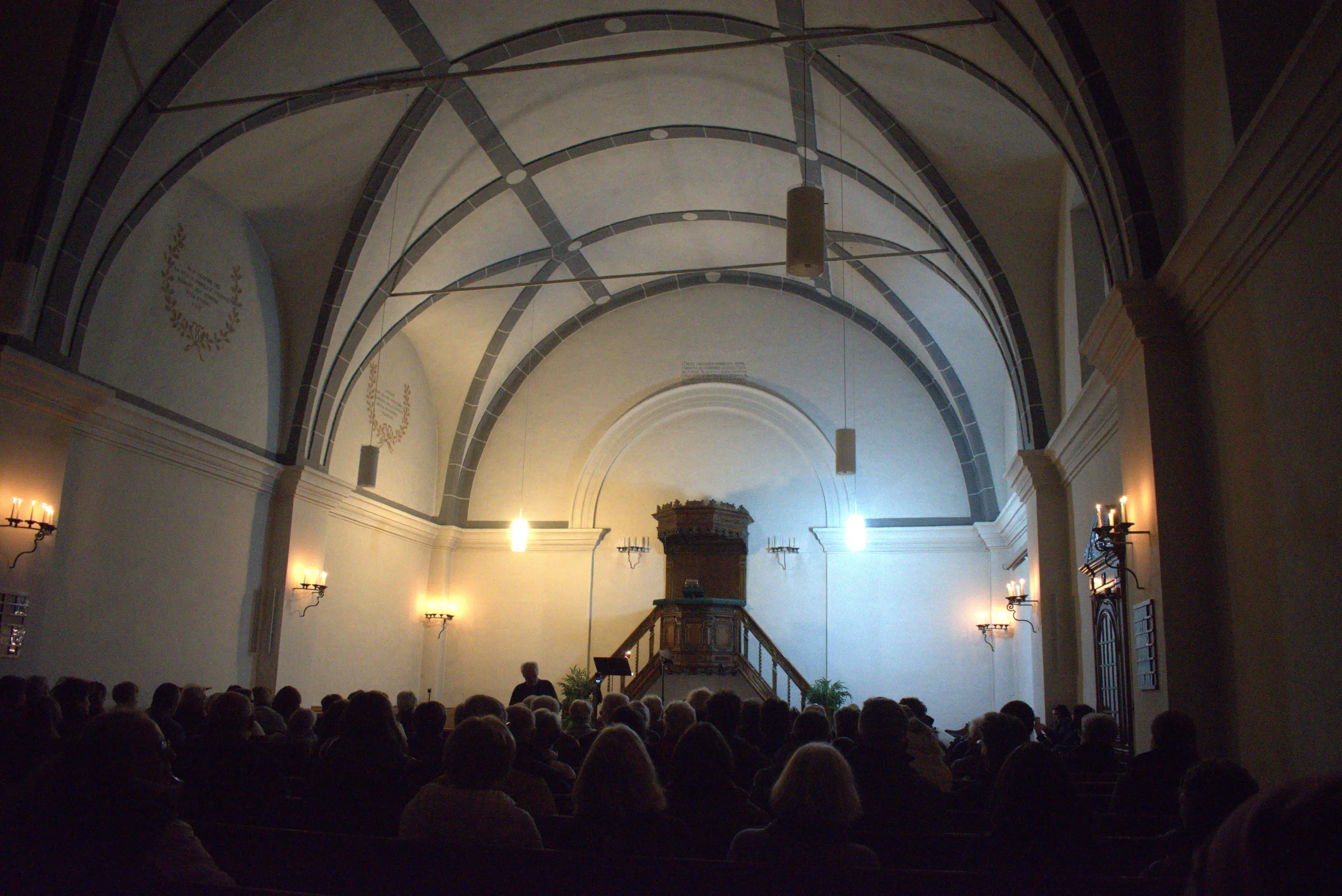 Una sala con un soffitto a volta illuminato. La scena mostra un gruppo di persone sedute di fronte a un grande pulpito in legno, con pareti chiare e eleganti lamp