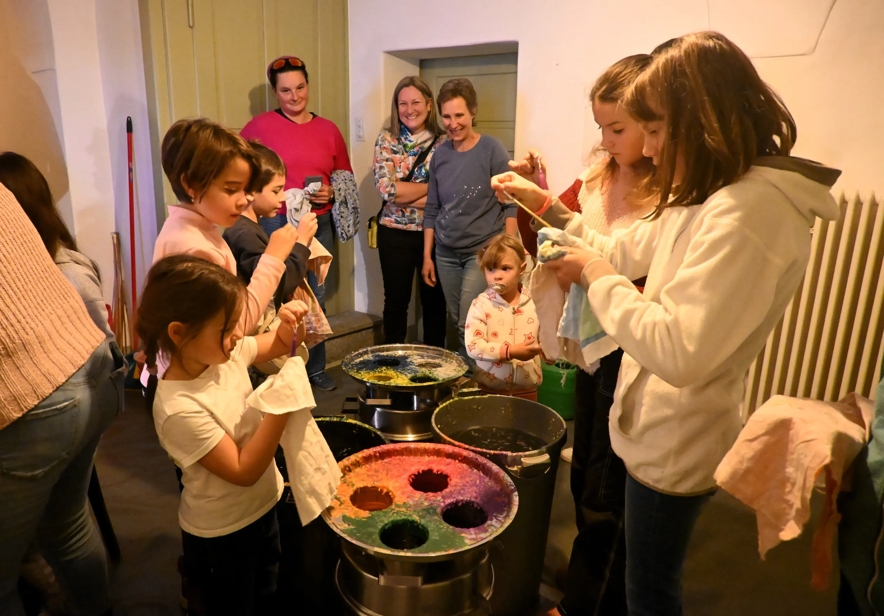 Un gruppo di bambini e adulti si riunisce in un ambiente interno, mentre i bambini sperimentano con colori in contenitori. Alcuni adulti osservano con interesse. C'è un'atmosfera di creatività e divertimento.