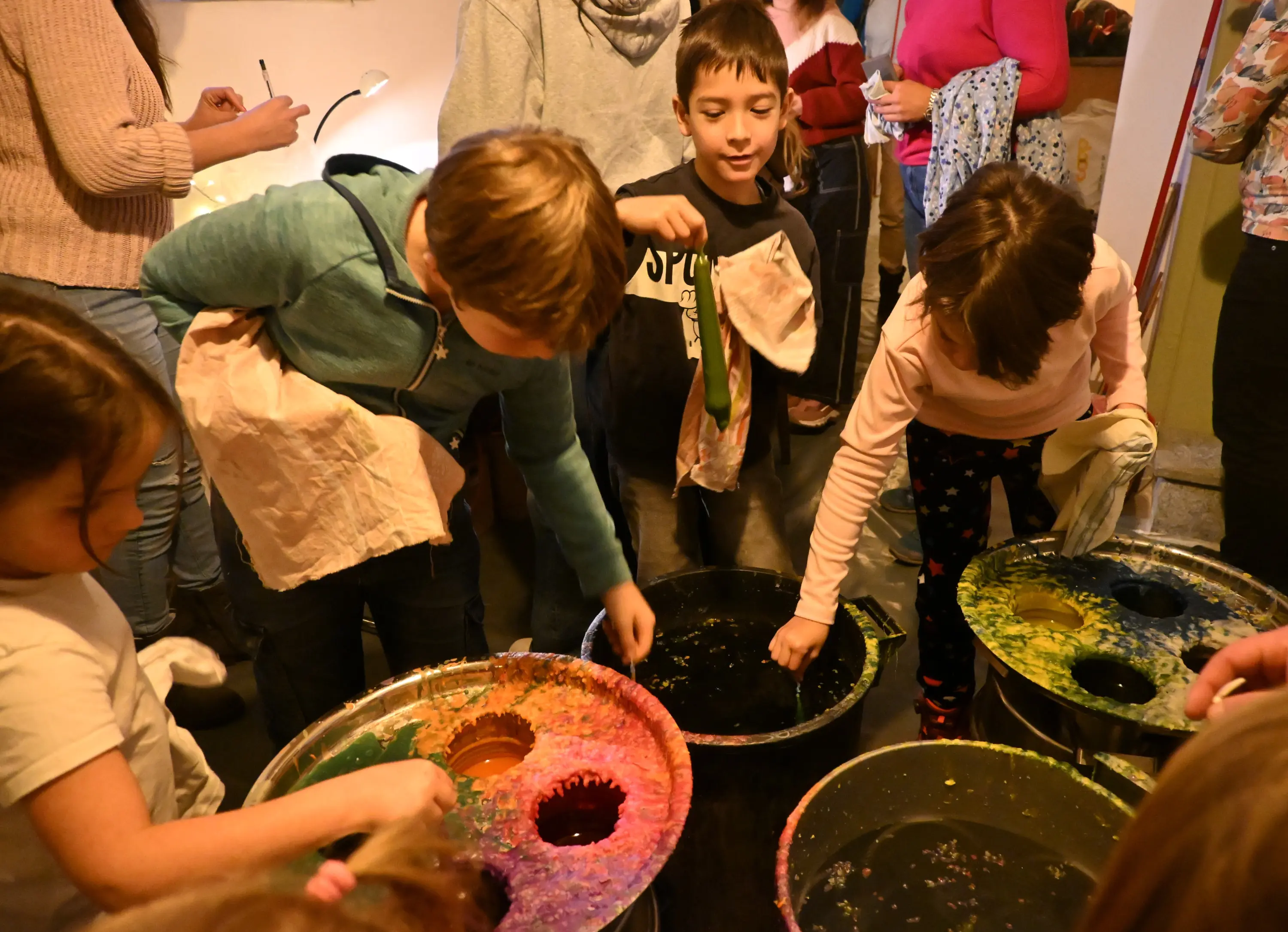 Gruppo di bambini che esplorano l'arte del colore, tuffando le mani in secchi di vernice multicolore. Alcuni indossano vestiti casual mentre si divertono in un ambiente creativo e vivace.