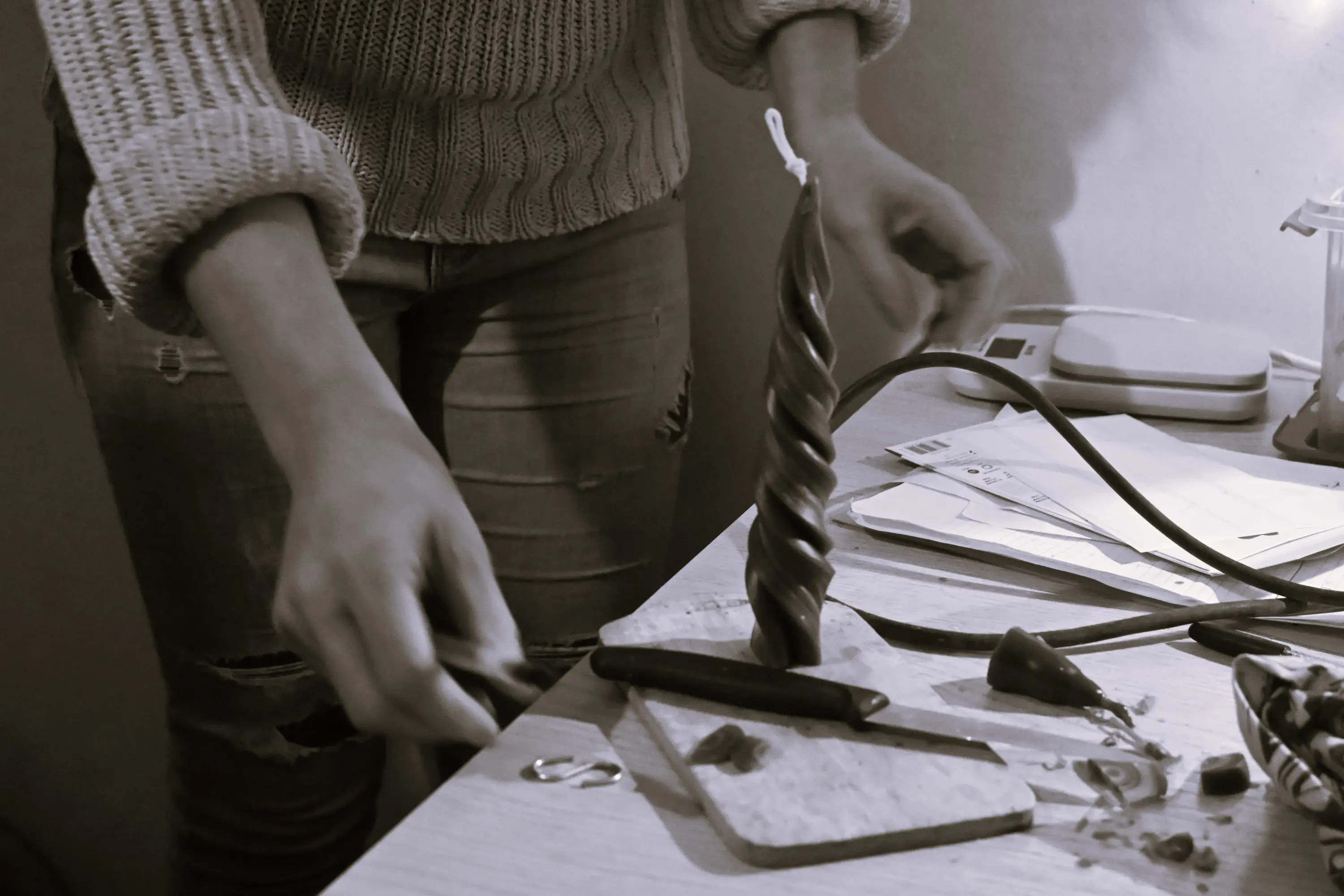 Una mano femminile tiene un piccolo oggetto mentre si trova vicino a una candela a spirale su un tavolo, circondata da attrezzi e materiali vari.