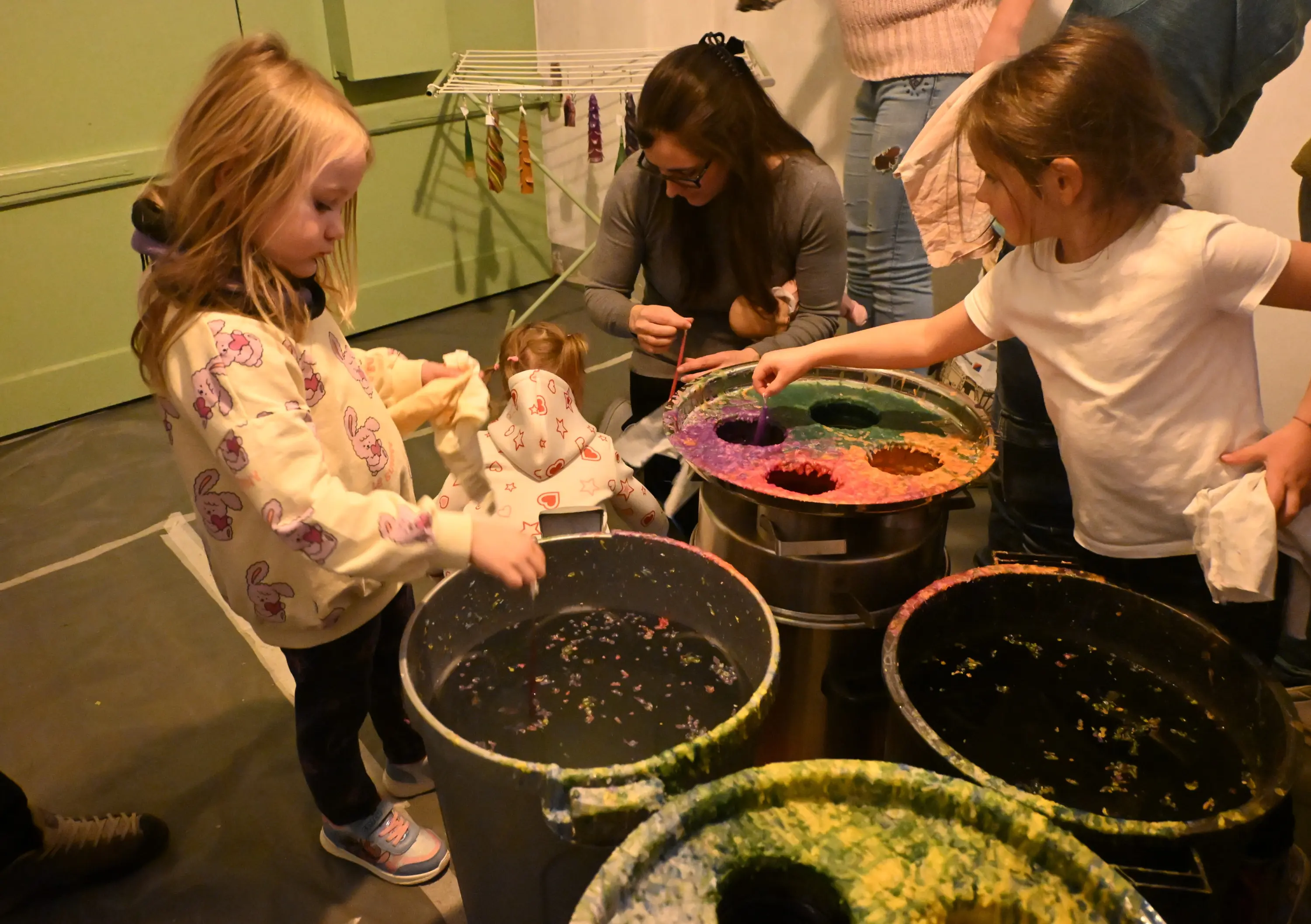 Due bambine stanno sperimentando con colori in grandi secchi. Una ragazza tinge un pezzo di tessuto, mentre l'altra osserva attentamente. Una donna adulta, seduta, le supervisiona. Altre persone si trovano sullo sfondo.