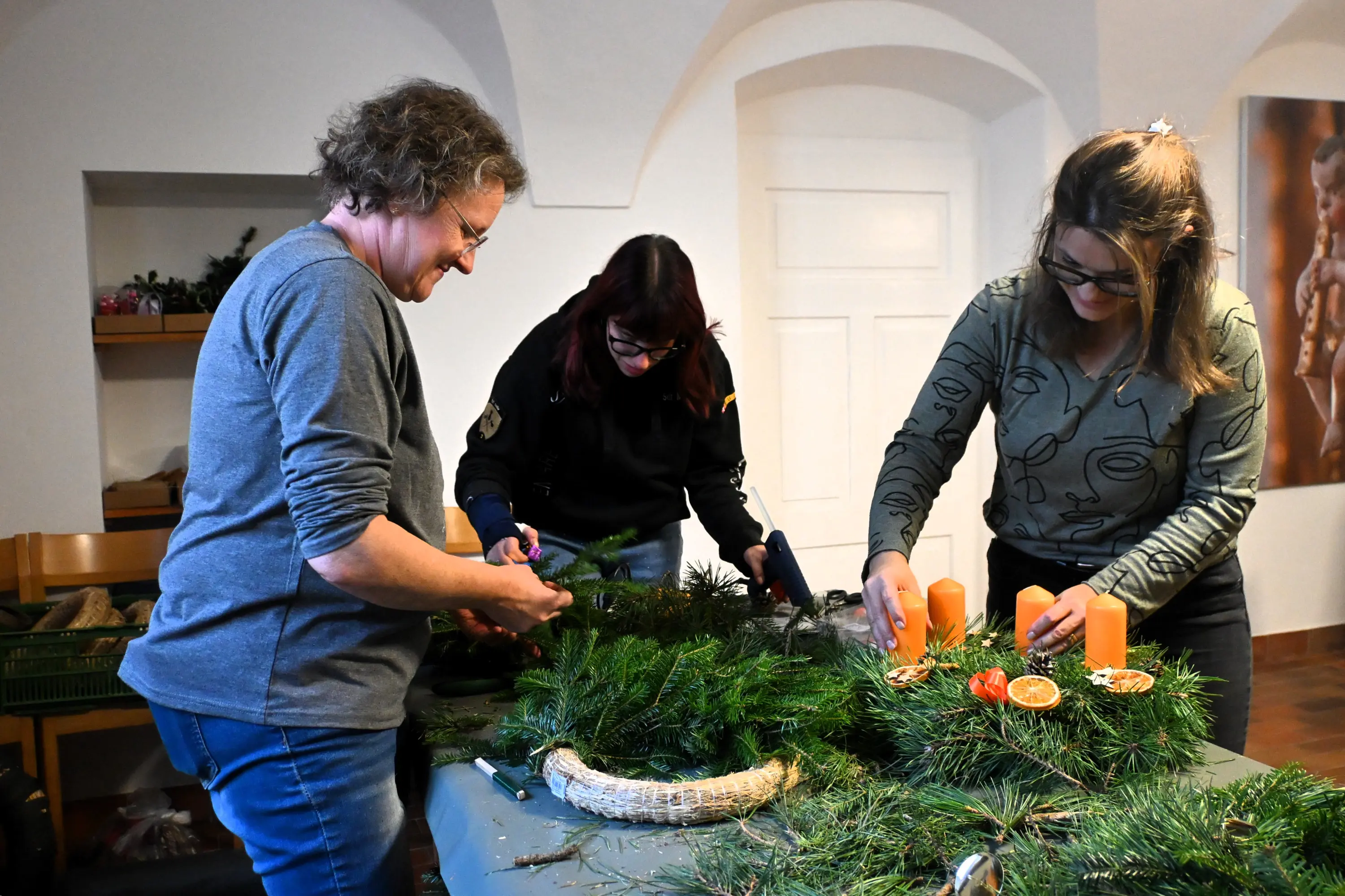 Tre donne creano decorazioni natalizie su un tavolo, utilizzando rami di pino, candele arancioni e elementi naturali. Tutte sono sorridenti e concentrate nel loro lavoro.