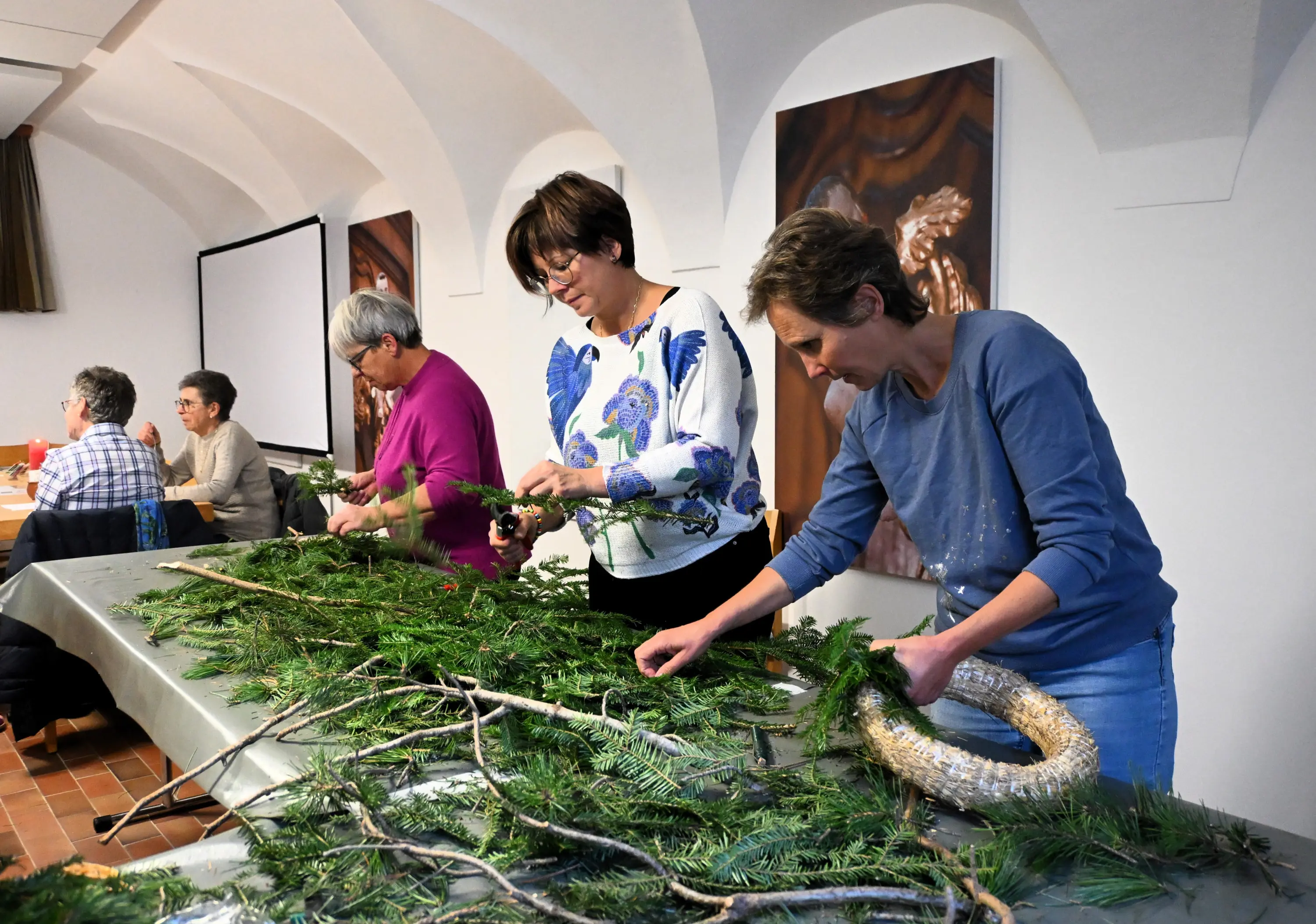 Tre donne stanno creando decorazioni natalizie con rami verdi e aghi di pino su un tavolo. Sono concentrate nel loro lavoro in un ambiente luminoso e accogliente. Sullo sfondo si vedono altre persone sedute e impegnate in attività simili.