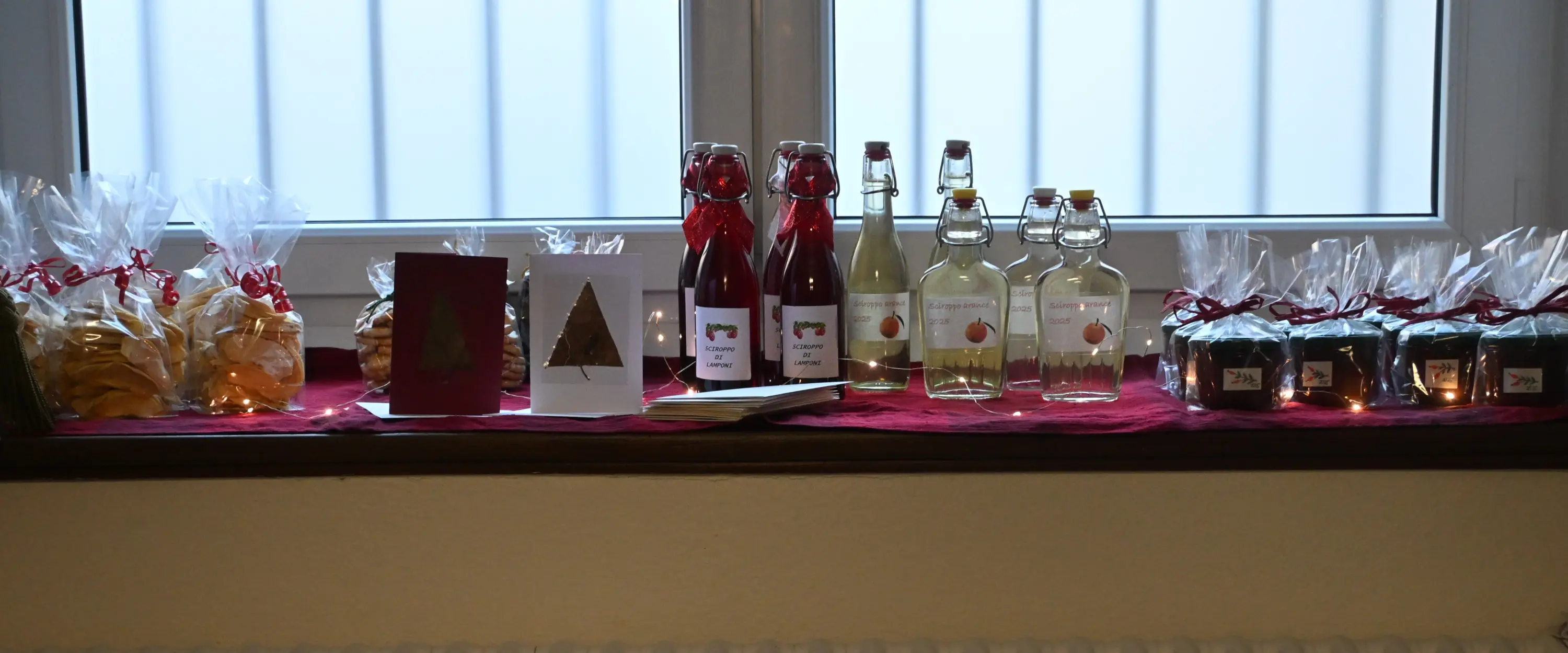 Una finestra con una varietà di prodotti artigianali su un davanzale. Ci sono bottiglie di liquori, marmellate e biscotti, tutti presentati con cura e decorati con nastri rossi. Una serie di candele accese aggiunge un'at