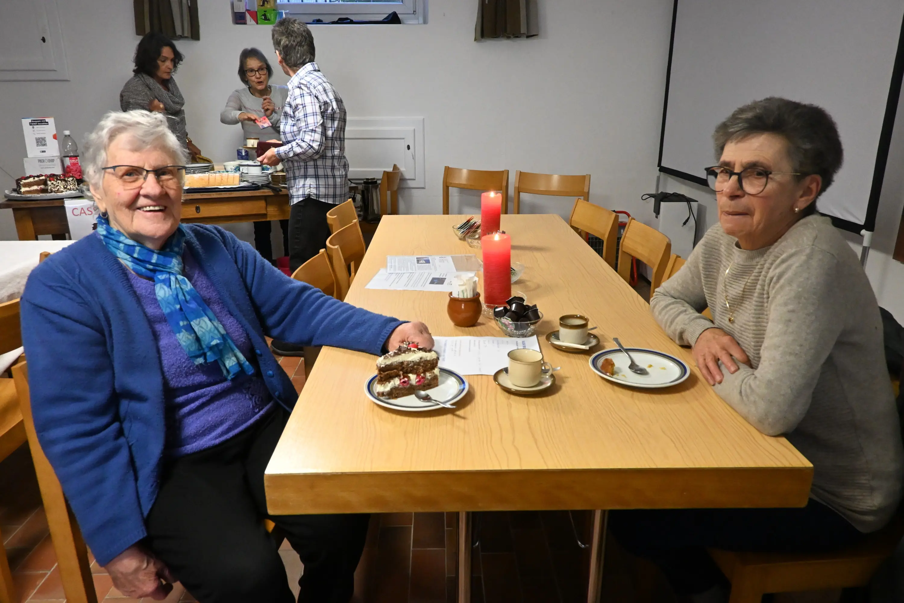 Due donne sedute a un tavolo di legno, sorridono mentre gustano dolci e caffè. Sul tavolo ci sono piatti con dessert, tazze e candele accese. Sullo sfondo, altre persone preparano cibo.