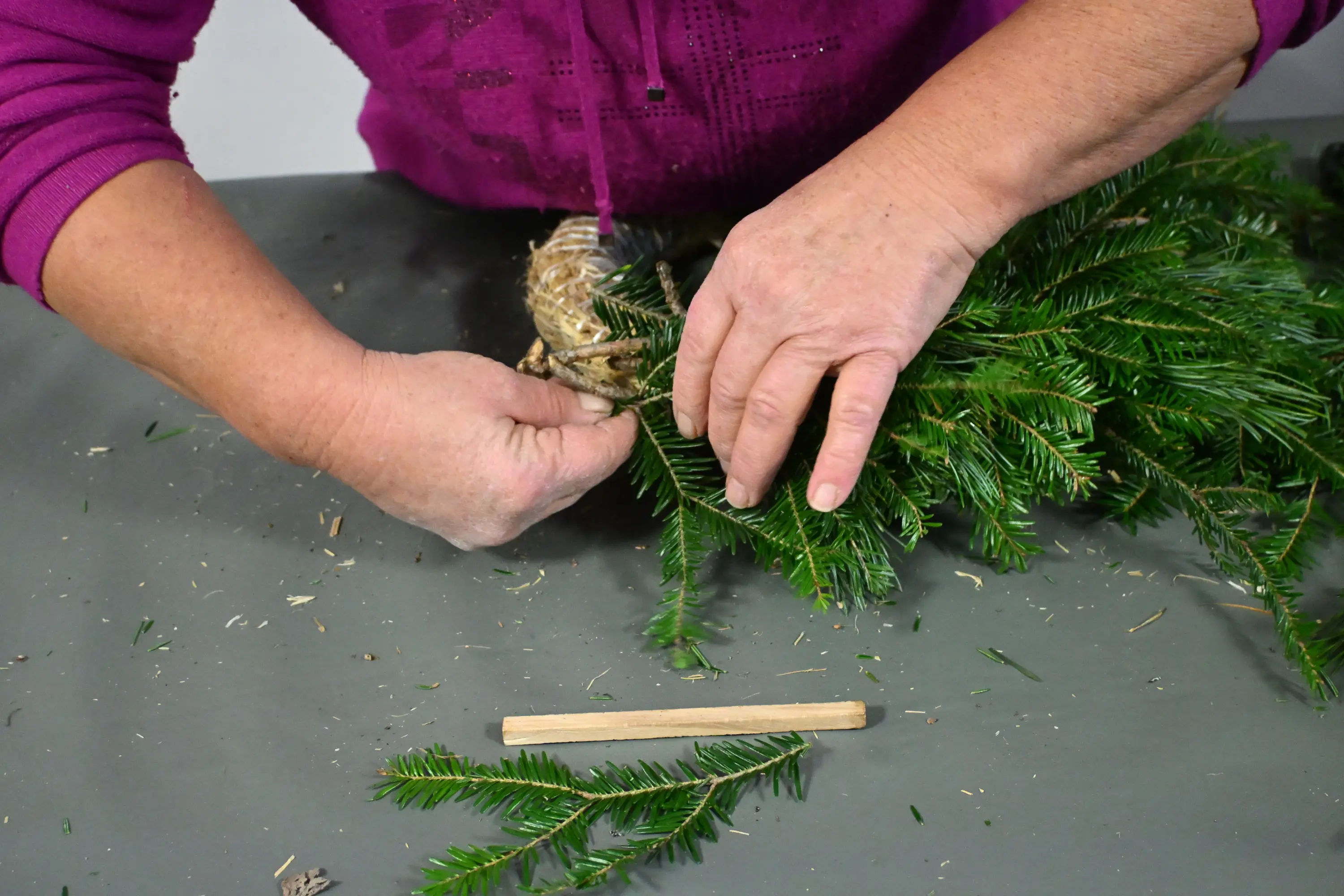 Mani che annodano rami di abete su un tavolo. Accanto, un bastoncino di legno e alcune foglie di pino sono sparsi.