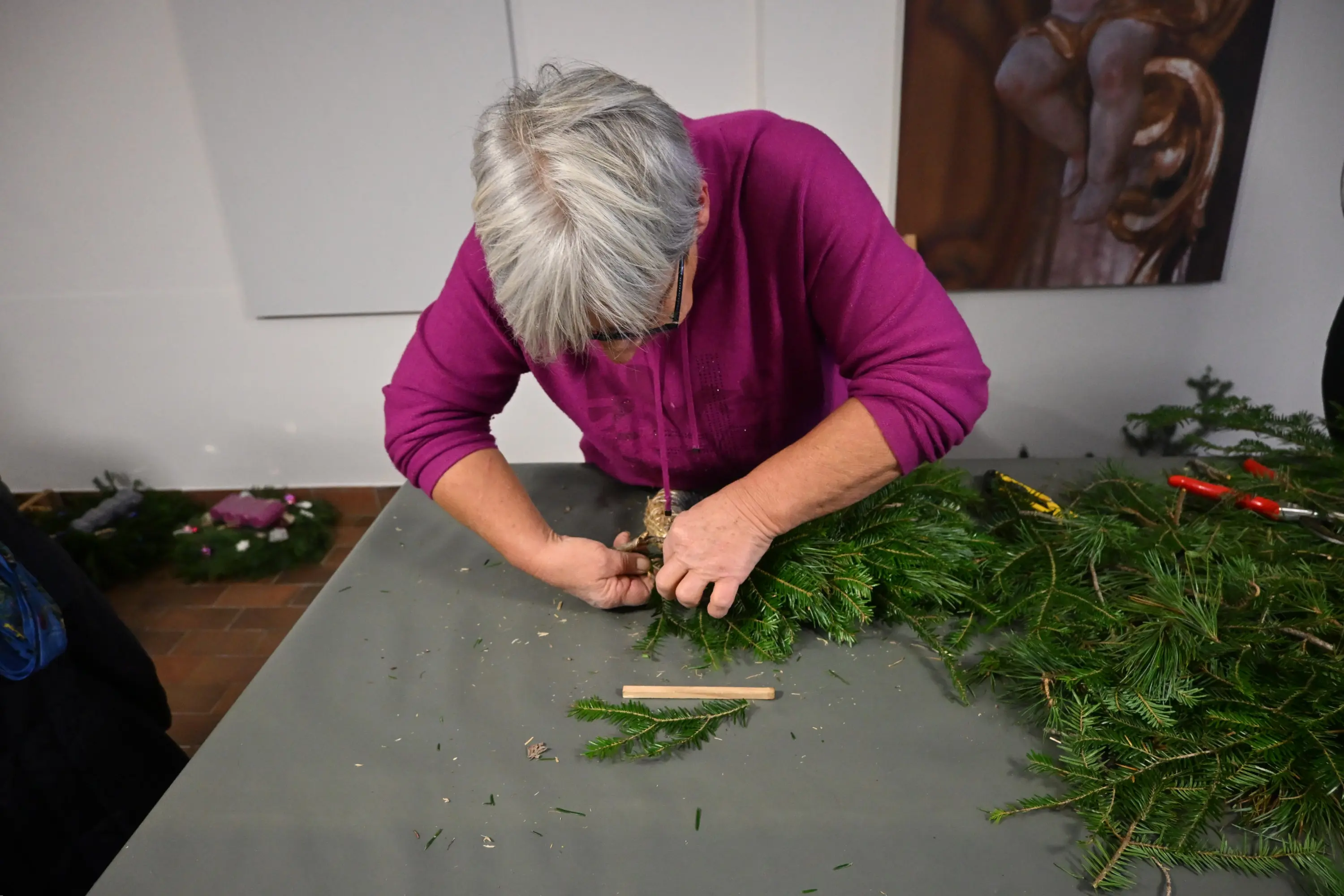 Una donna con i capelli grigi sta creando un decorazione natalizia usando rami di abete su un tavolo. Indossa una maglia viola e appare concentrata nel suo lavoro.